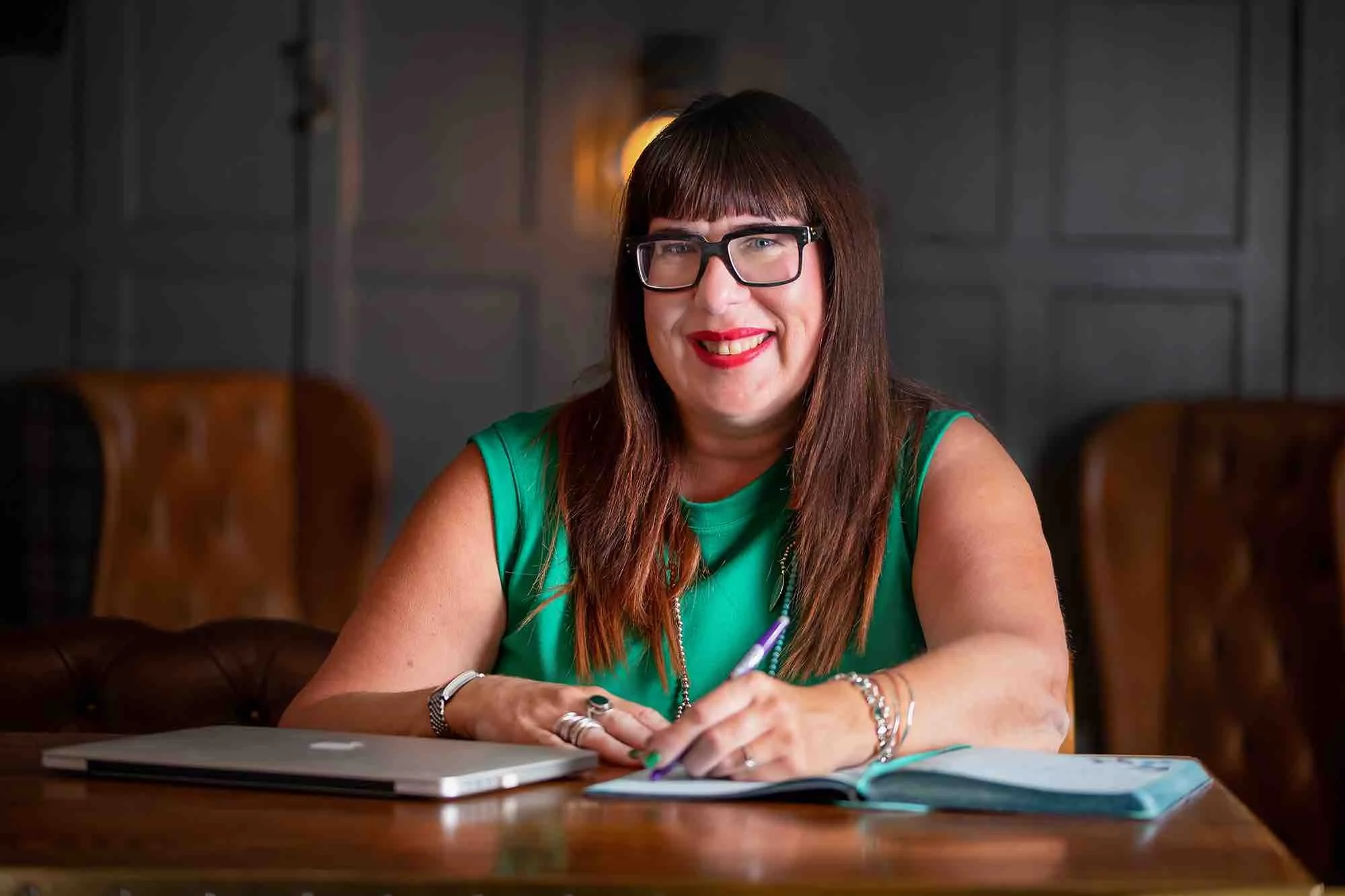 A woman sitting at a table with a laptop, writing in a notebook, smiling, wearing glasses and a green top in a warmly lit room.