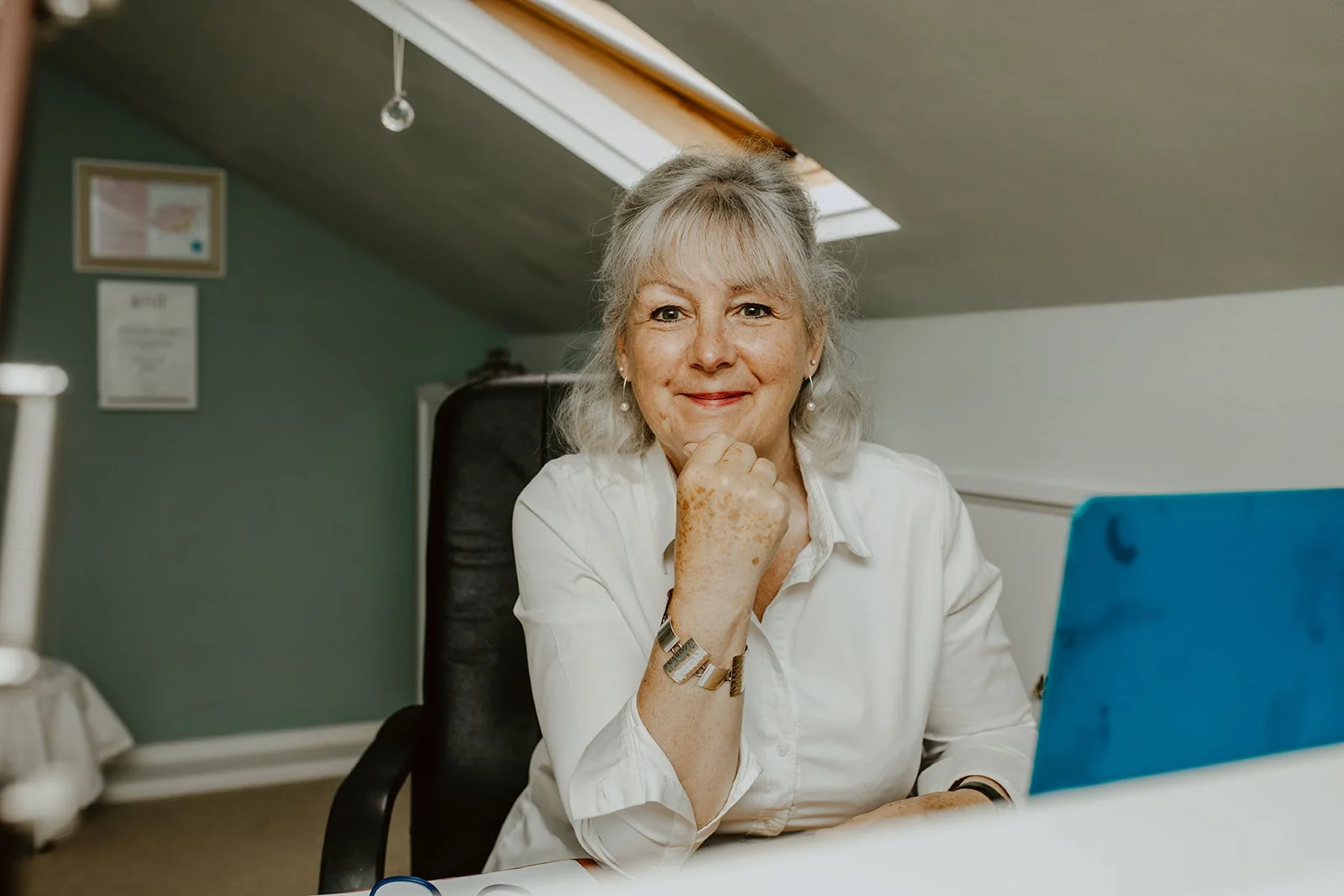 A smiling woman sitting at a desk in an office or home workspace, looking at the camera with her hand resting under her chin.