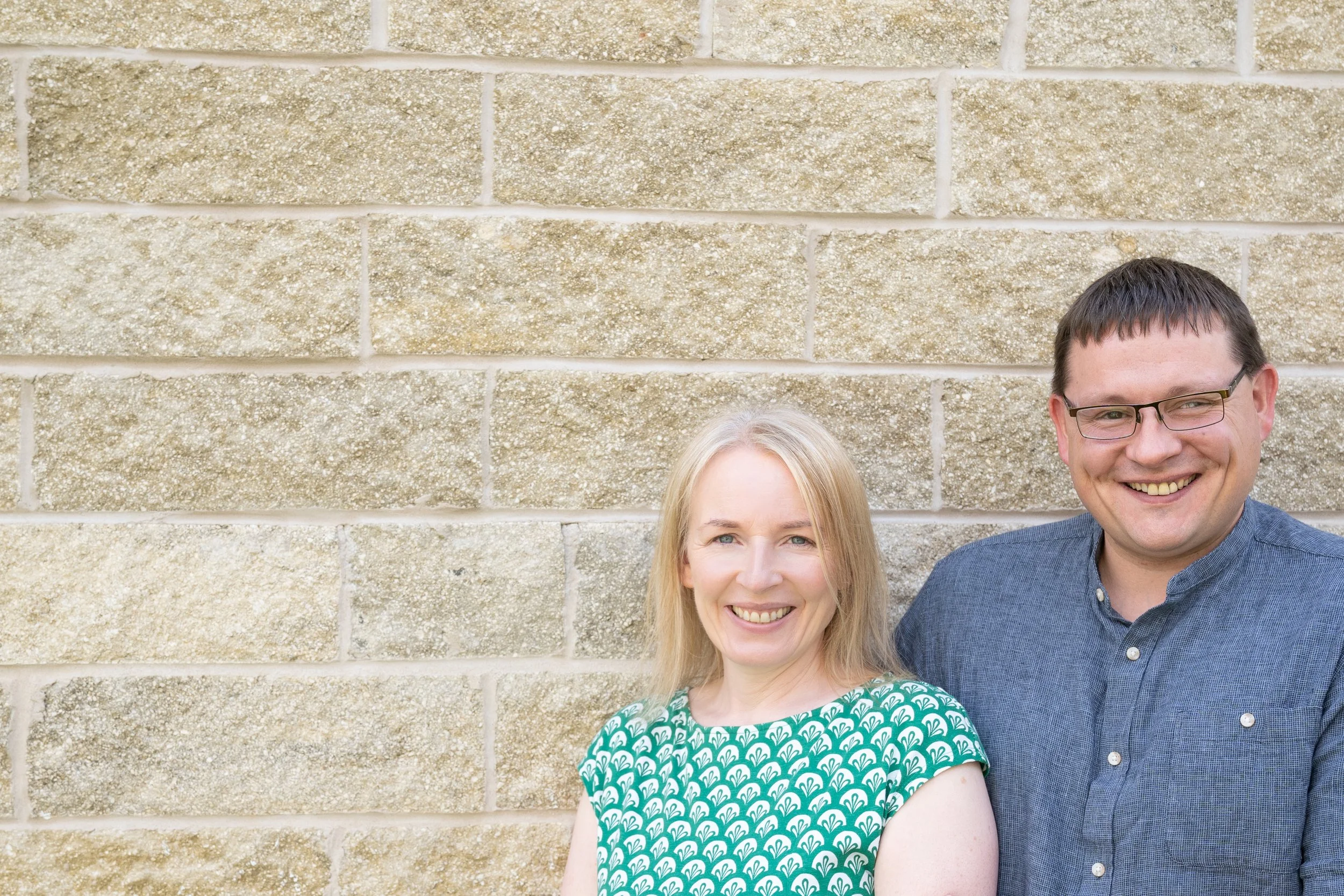 A smiling woman with blonde hair wearing a green patterned dress standing next to a smiling man with glasses and dark hair wearing a blue button-up shirt, standing against a beige brick wall.