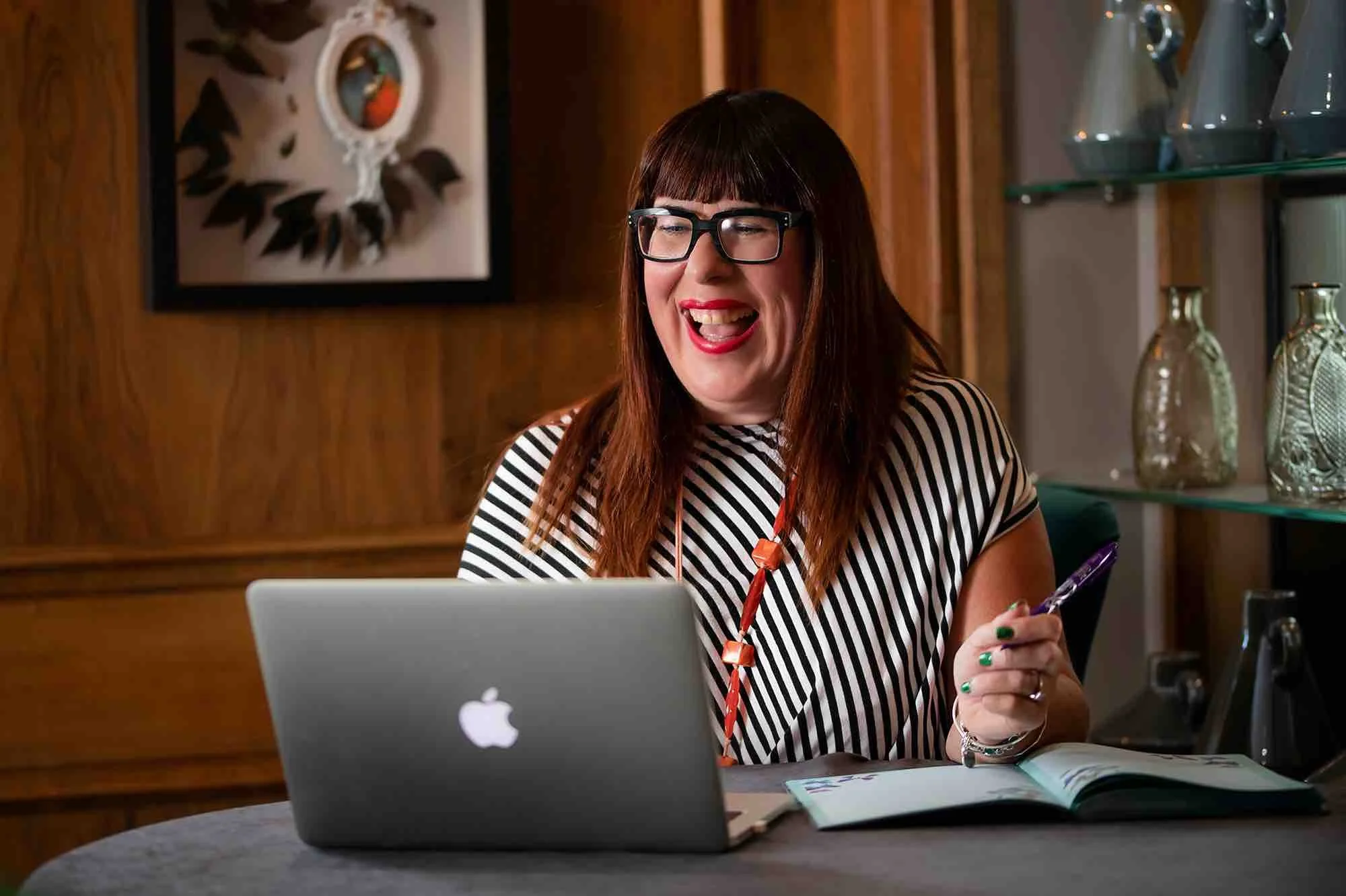 A woman with glasses and a striped shirt sits at a desk, smiling and talking while working on a laptop. An open notebook and a pen are in front of her, and there are decorative glass items on the shelves behind her.