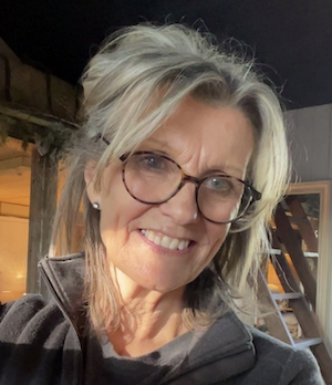 A woman with glasses and blonde hair smiling at the camera indoors.