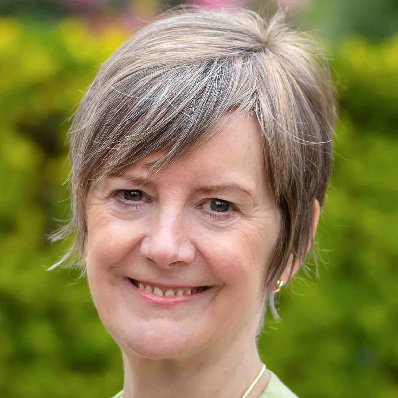 Close-up portrait of a smiling woman with short gray hair, wearing a gold hoop earring and a necklace, outdoors with blurred green foliage in the background.