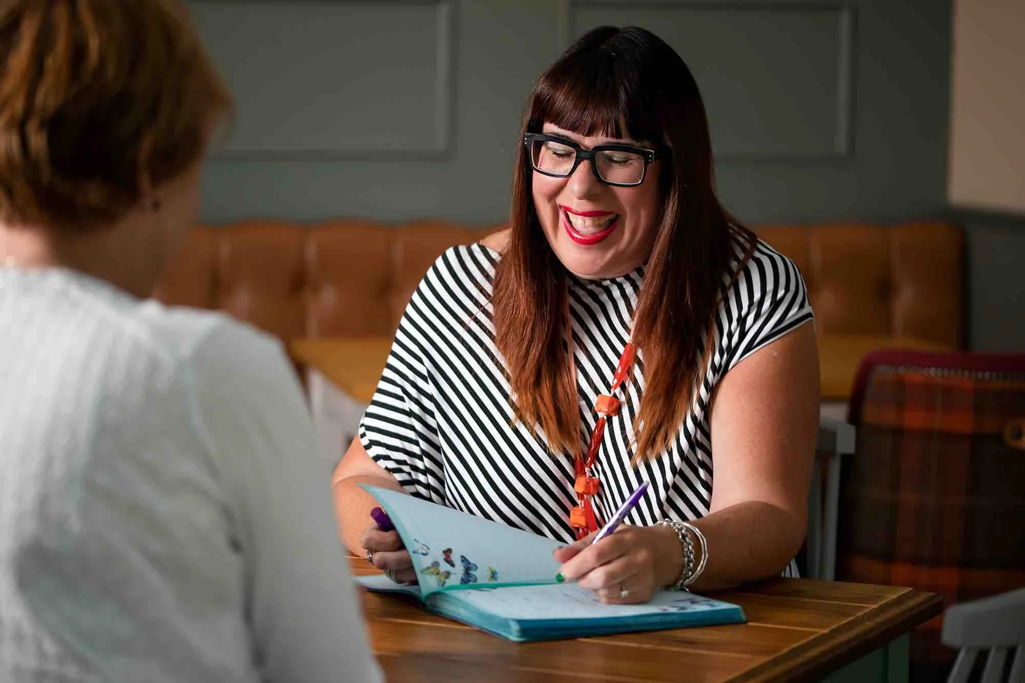 A woman with long brown hair, wearing glasses and a black-and-white striped shirt, smiling while holding a pen, sitting at a wooden table during a conversation with another person.