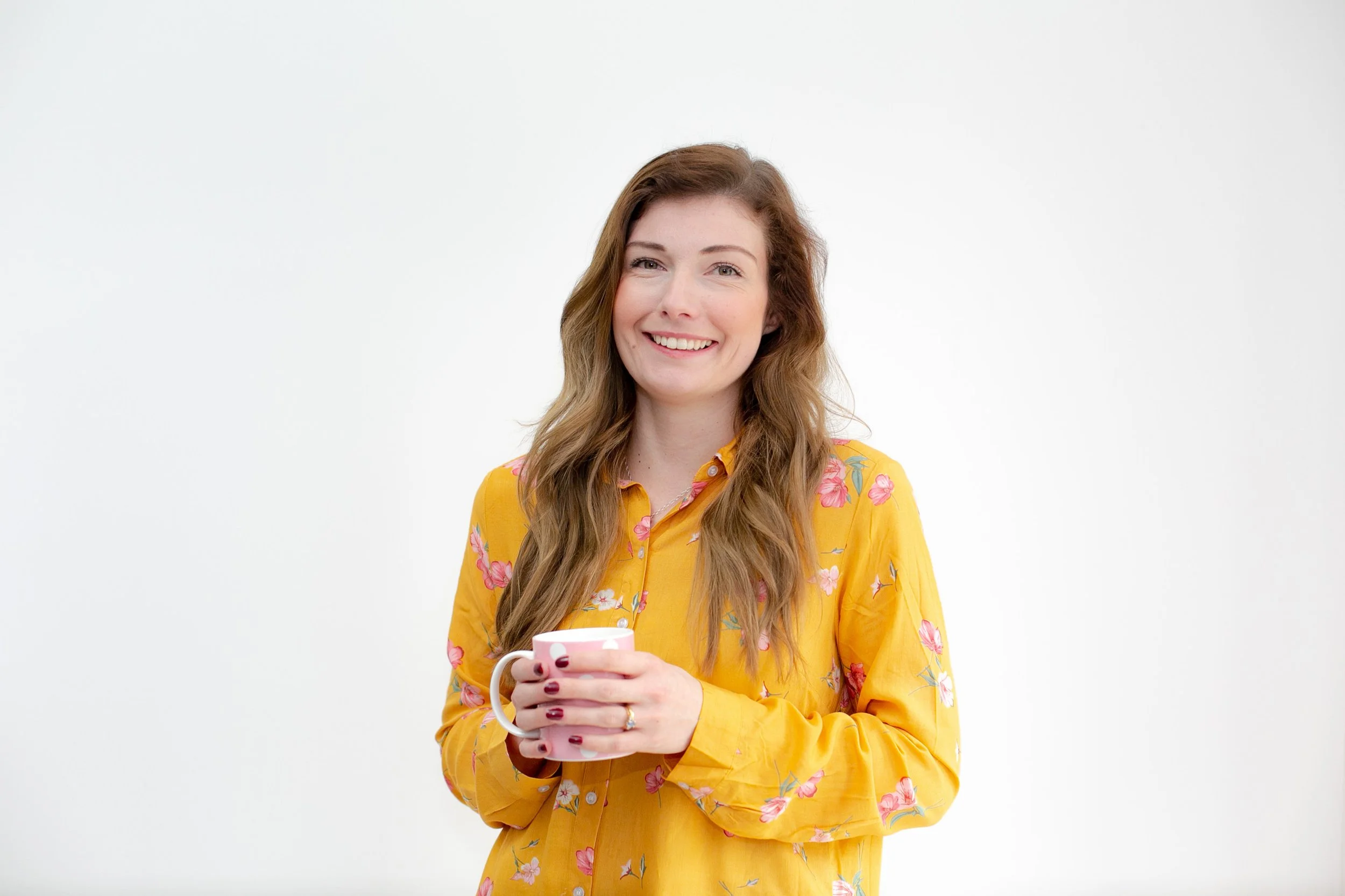 A smiling woman with long wavy hair holding a white mug with a pink design, wearing a yellow floral shirt, standing against a plain white background.