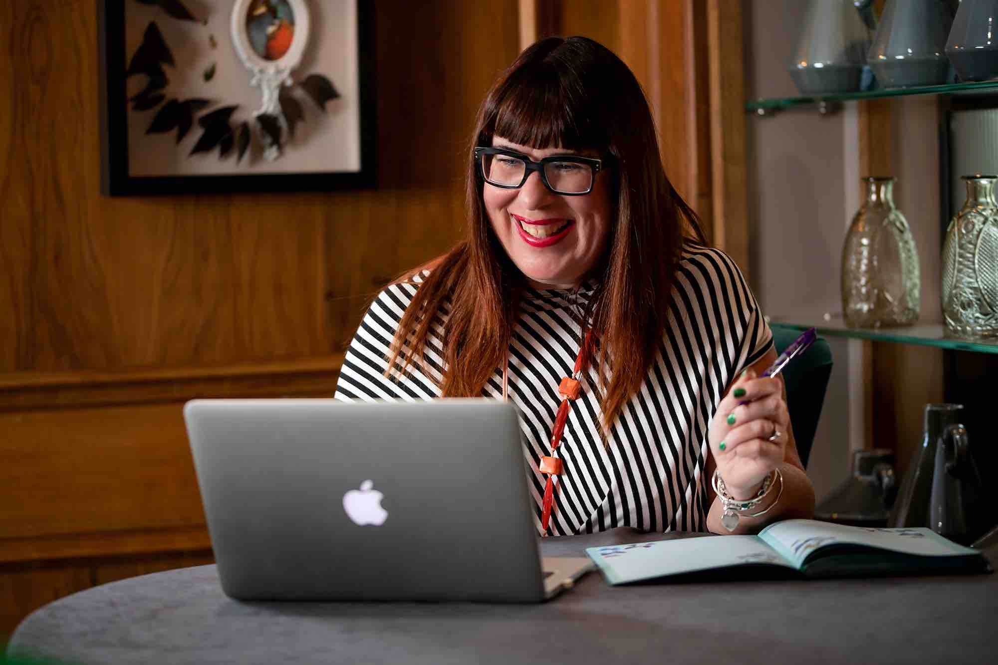 Karen Taylor, website creator sitting at a table with a silver MacBook, open books, and holding a pen in a cozy wooden interior.