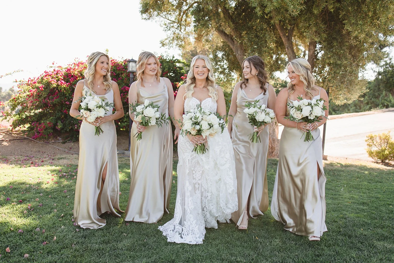 Bride in a white lace wedding gown walking with five bridesmaids in champagne colored dresses outdoors under a large tree, holding white floral bouquets.