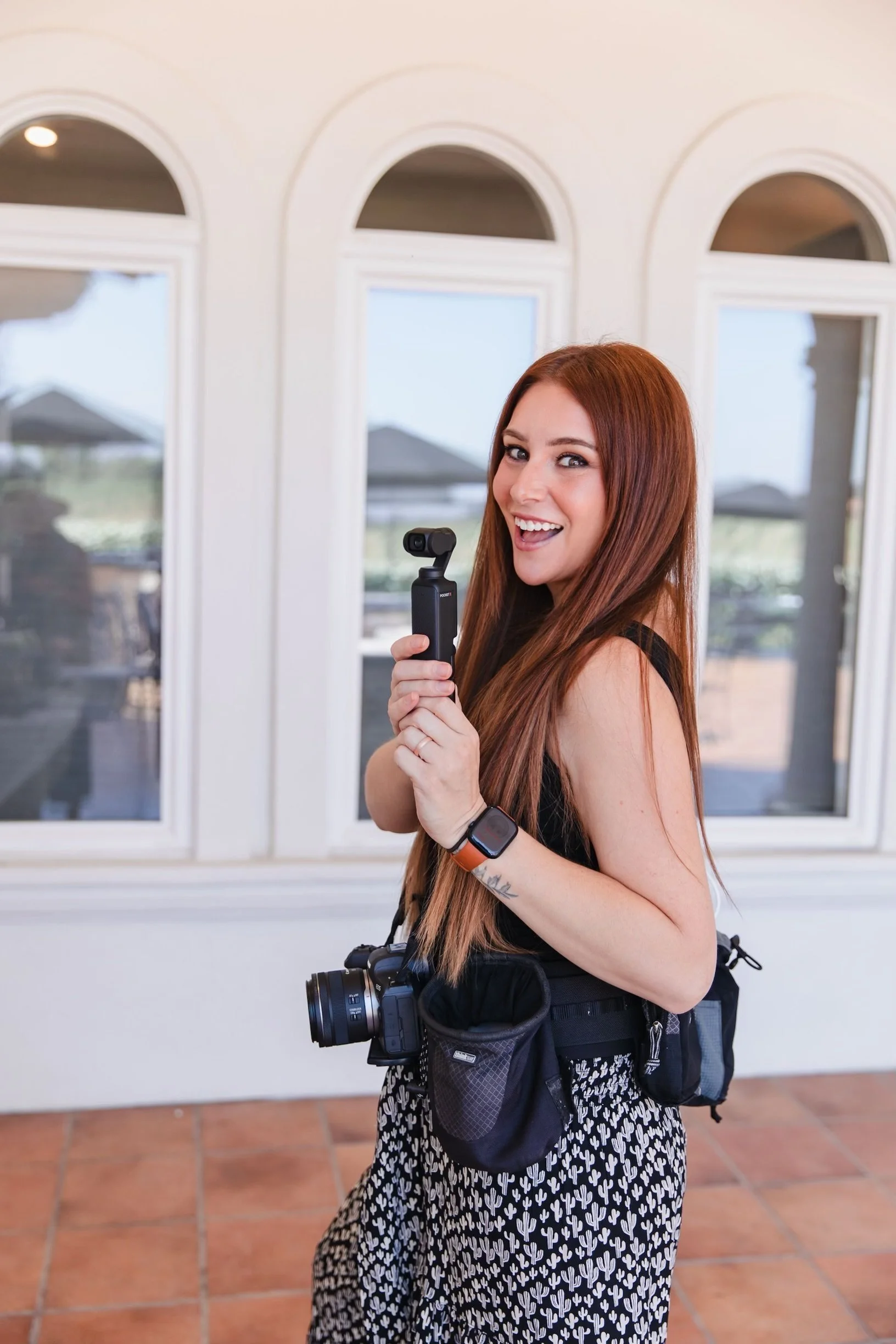 A woman with long red hair smiling and posing with a camera and a gimbal, indoors in front of arched windows.