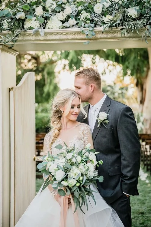 Bride and groom sharing a moment under an archway decorated with flowers, outdoors in a garden setting.