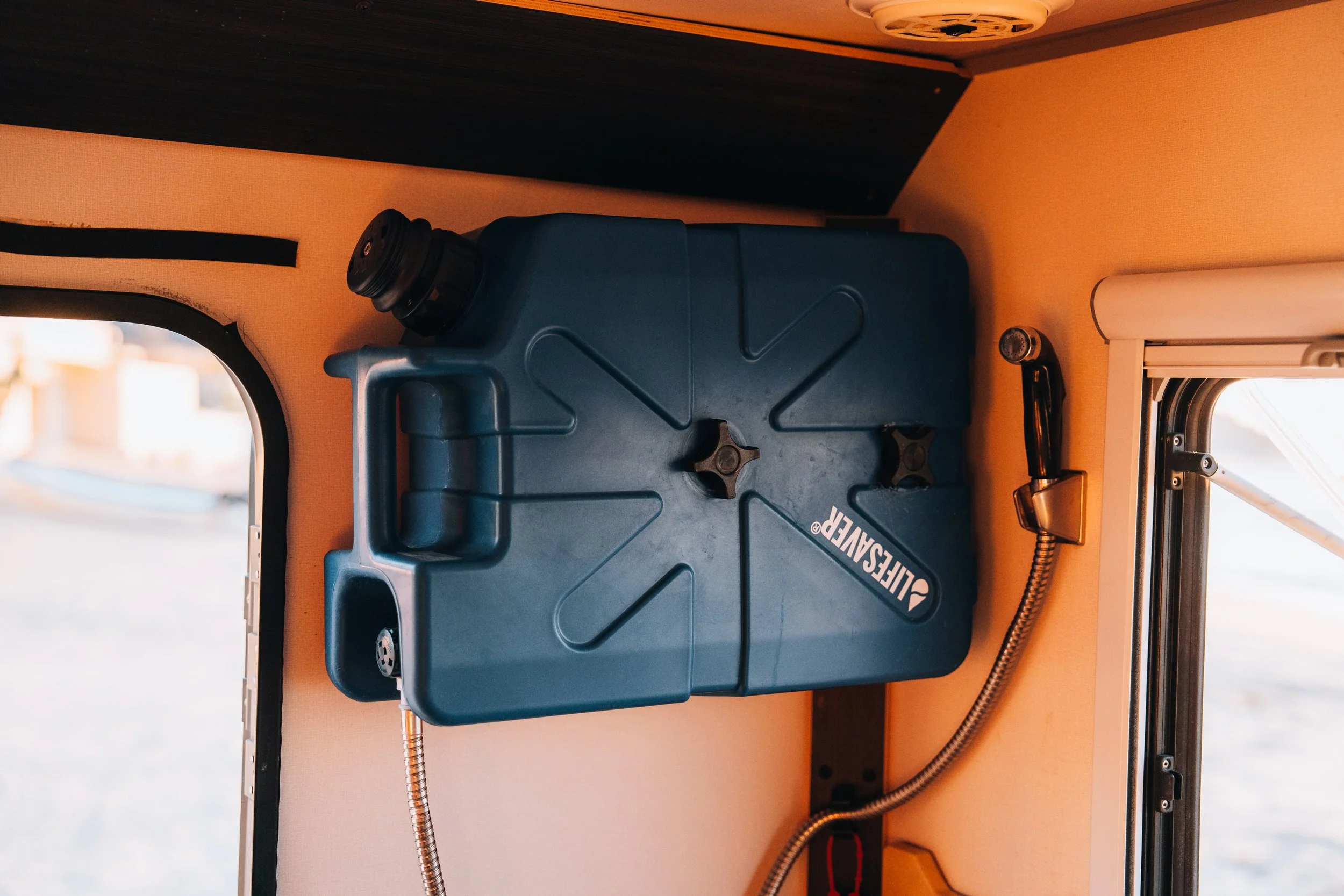 Blue Diversaew plastic container with a black cap, a vent, and a flexible black and silver hose, placed on a beige surface, with part of a window and window frame visible in the background.