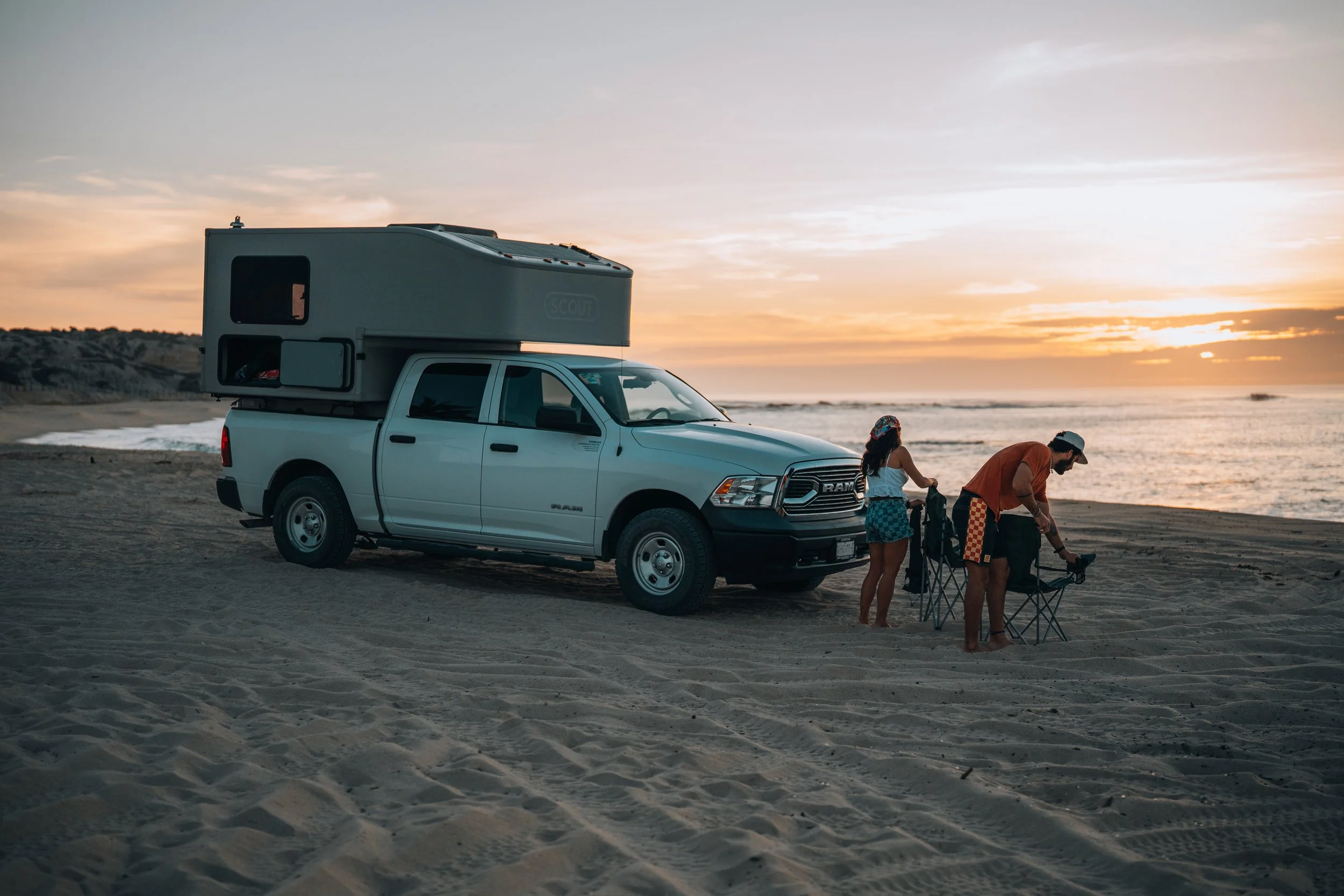 People enjoying a day at the beach with an RV, vehicle, camping chairs, and a table with drinks and supplies on sandy beach under partly cloudy sky.