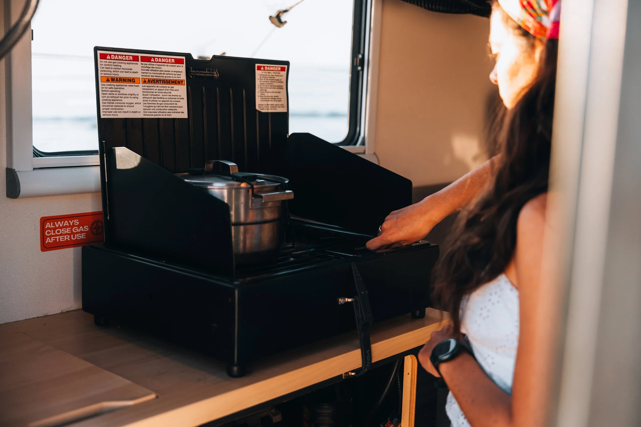 Small kitchen area inside a camper or RV with a Dometic electric mini fridge and a black AC/DC cooler on a light wood countertop, sign reminding to close gas after use, and a small cabinet underneath containing a spray can and other items.