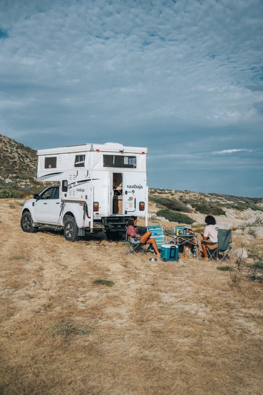 People enjoying a day at the beach with an RV, vehicle, camping chairs, and a table with drinks and supplies on sandy beach under partly cloudy sky.