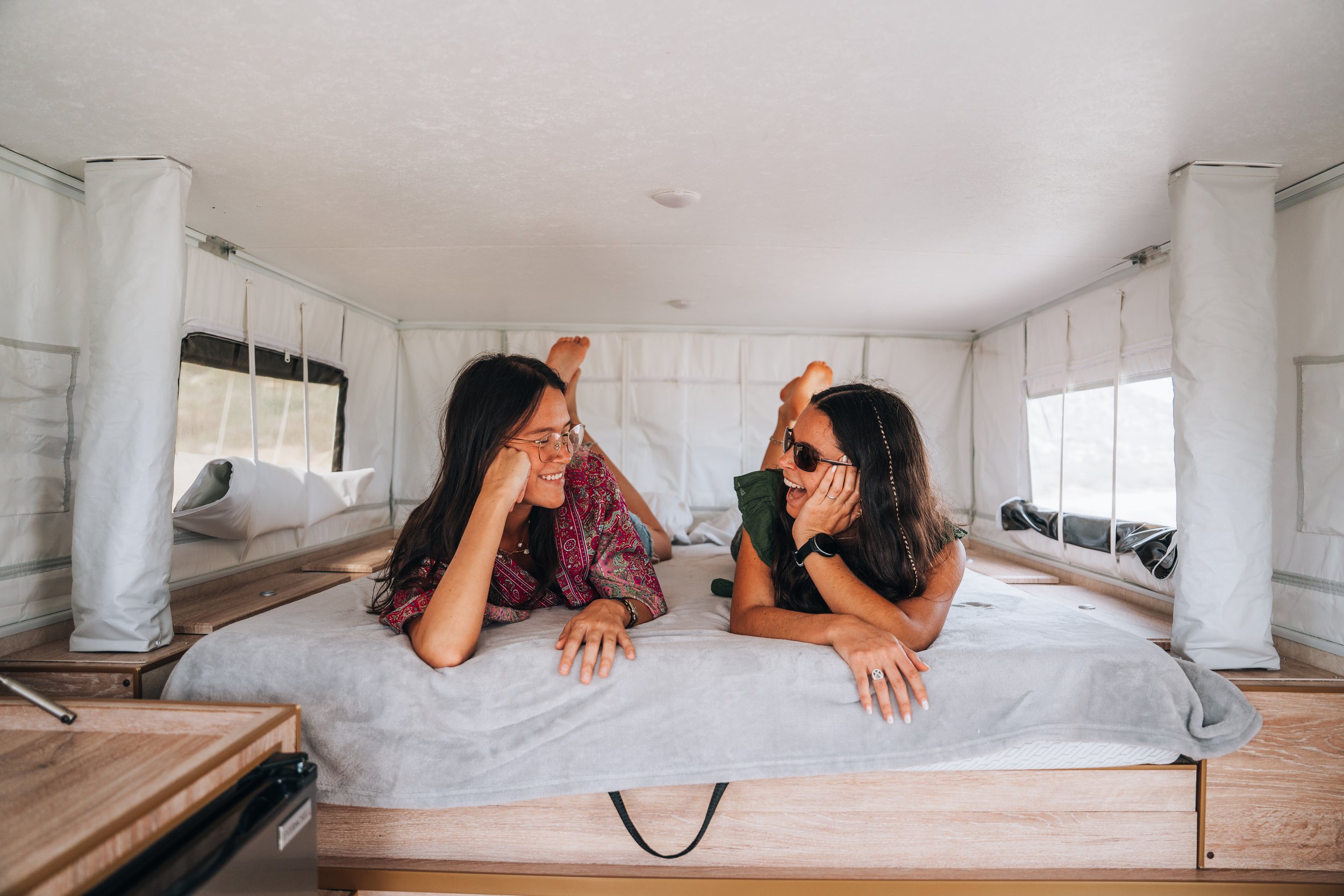 Ad woman lying on her side on a bed, resting her head on a pillow, with an orange knit blanket and white sheets, inside an RV or camper with a skylight overhead.