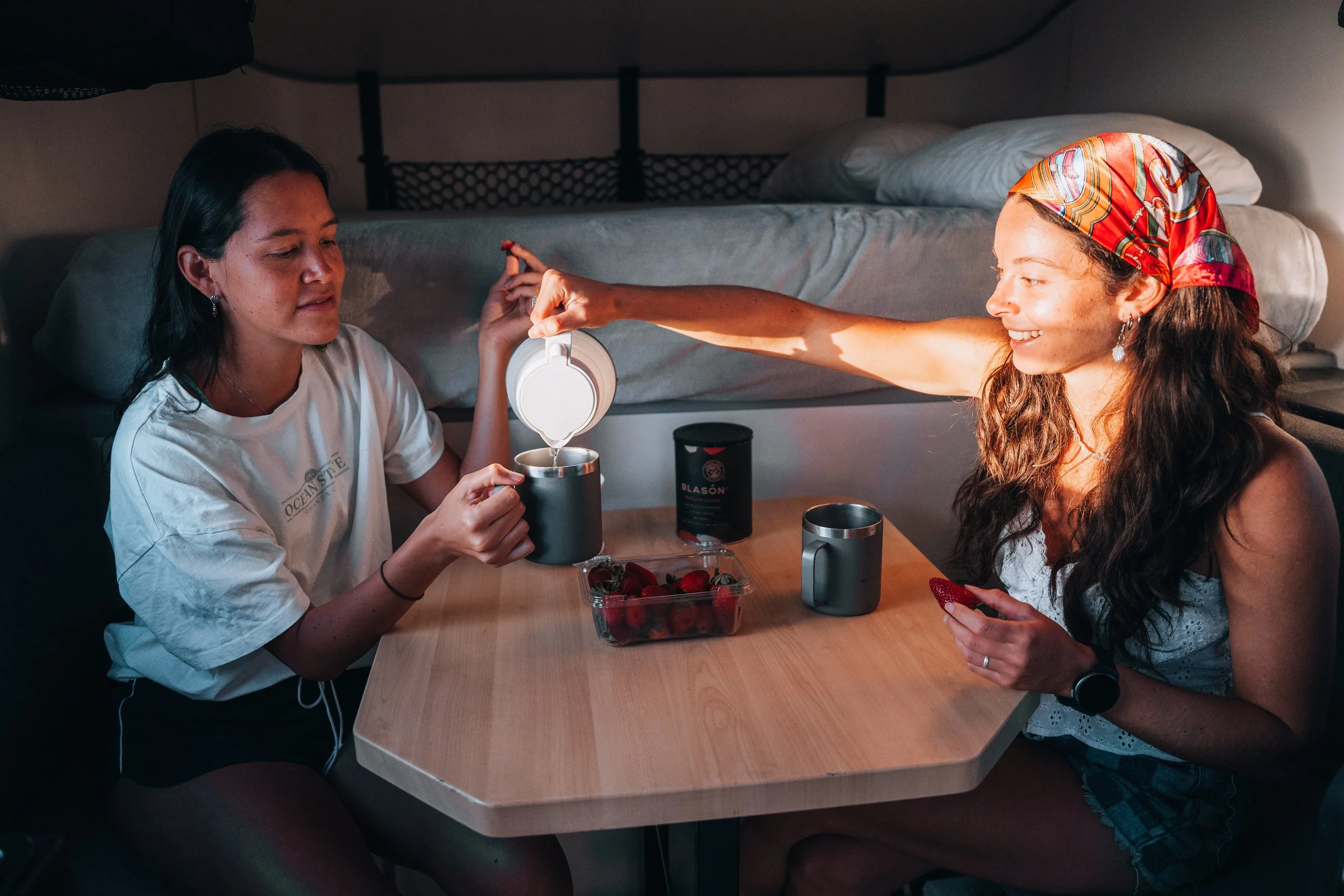 A wooden dining table with black metal legs is positioned between two gray cushioned benches. In the background, there is a bed with a white sheet and an orange knitted blanket folded at the foot of the bed.