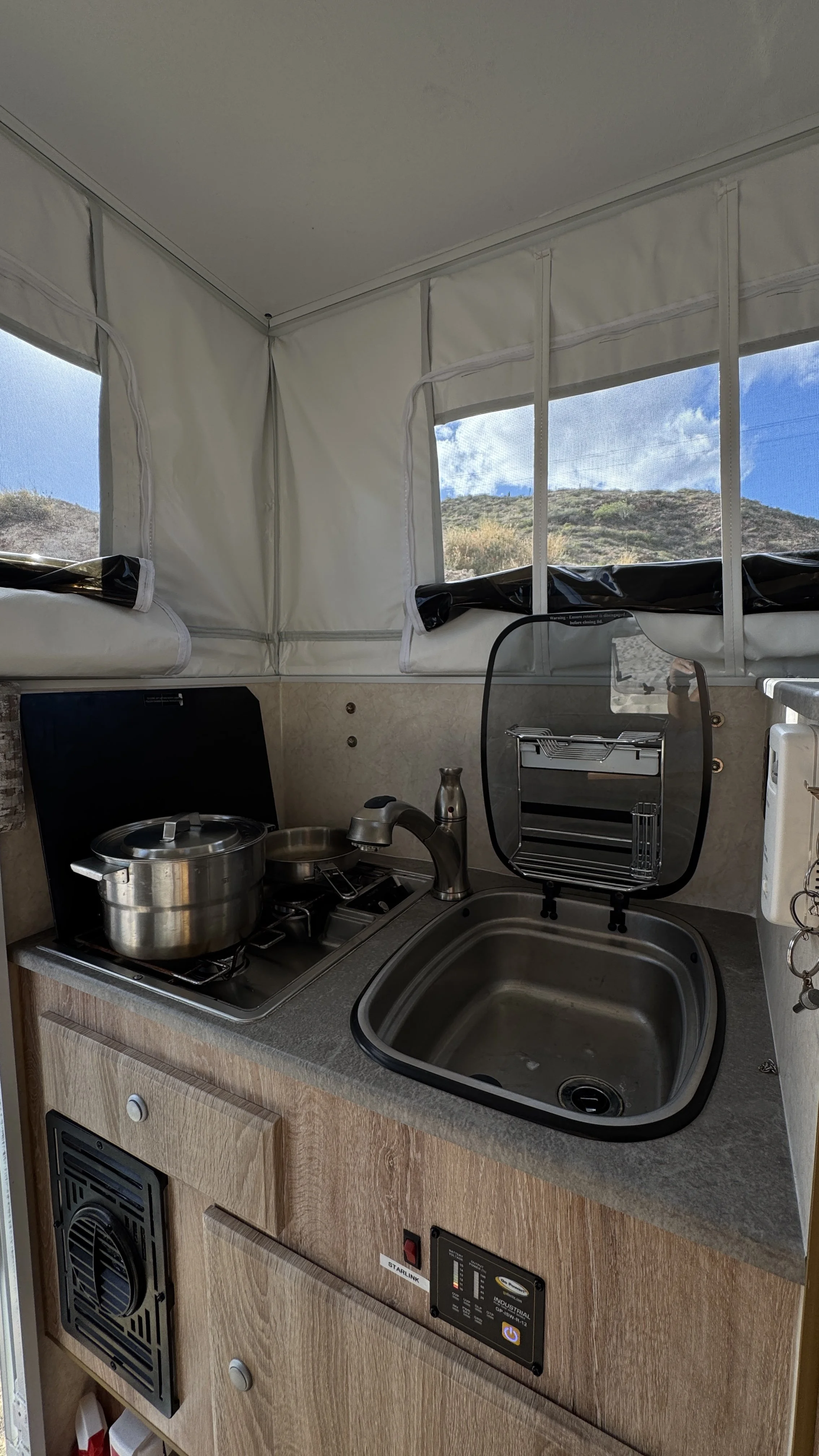 Small kitchen area inside a camper or RV with a Dometic electric mini fridge and a black AC/DC cooler on a light wood countertop, sign reminding to close gas after use, and a small cabinet underneath containing a spray can and other items.