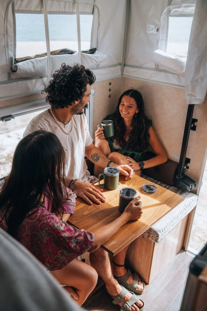 A wooden dining table with black metal legs is positioned between two gray cushioned benches. In the background, there is a bed with a white sheet and an orange knitted blanket folded at the foot of the bed.