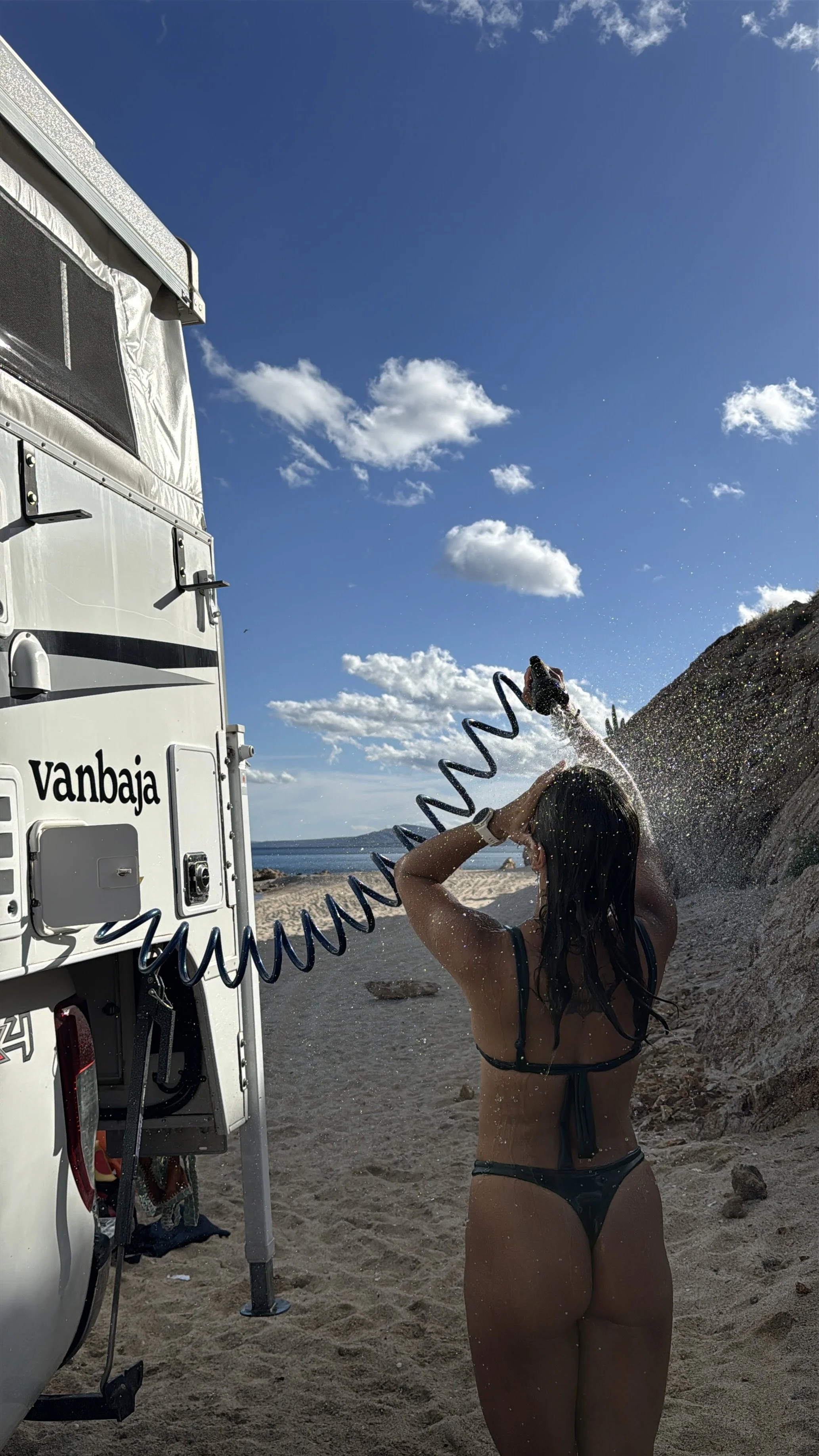 Woman in a bikini rinsing off with water at the beach next to a campervan with the signage 'vanbaja'.