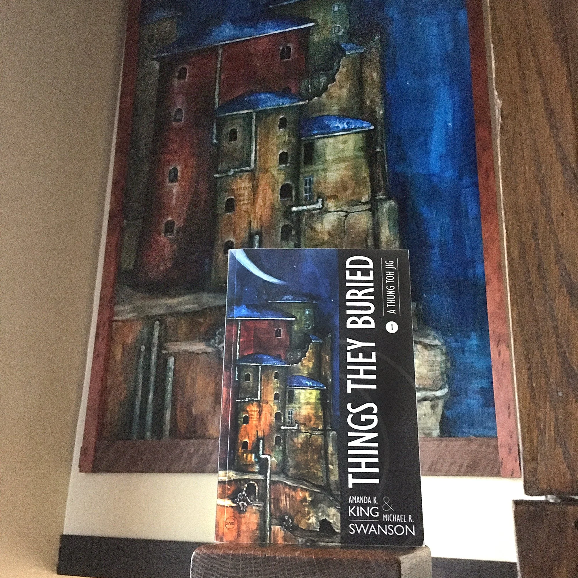 Indie published novel, Things They Buried by Amanda K King & Michael R Swanson, held in front of the cover painting by Michael B Fee