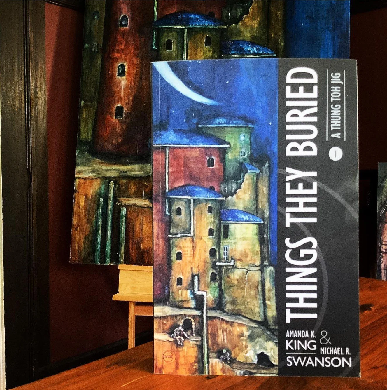 Indie published novel, Things They Buried by Amanda K King & Michael R Swanson, sitting in front of the cover painting by Michael B Fee