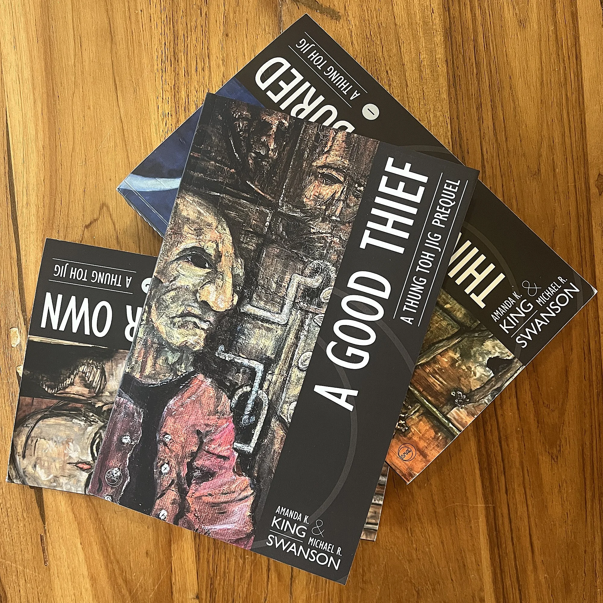A Good Thief novella trade paperback lying on top of series novels Things They Buried and They Eat Their Own