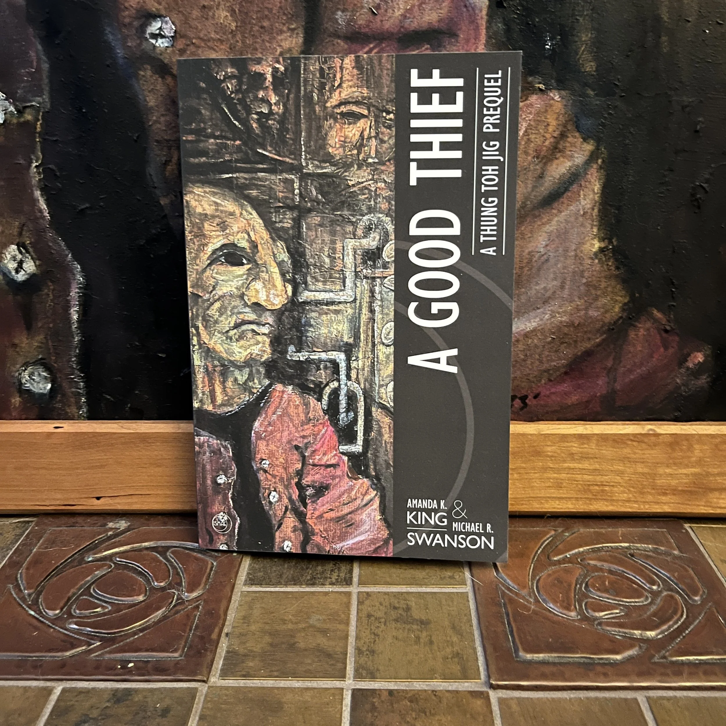 A Good Thief novella by Amanda K .King and Michael R. Swanson sitting in front of the cover painting by Michael B. Fee