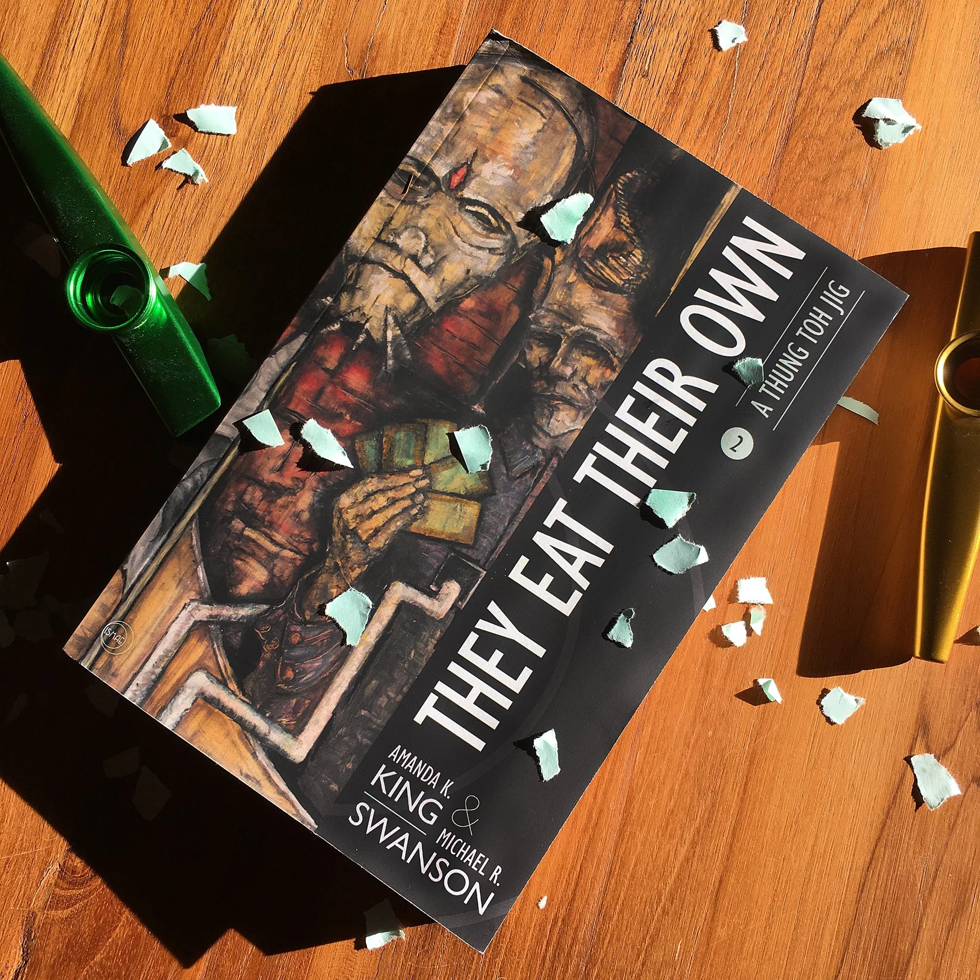 They Eat Their Own trade paperback surrounded by scattered confetti, curly ribbons, and a kazoo