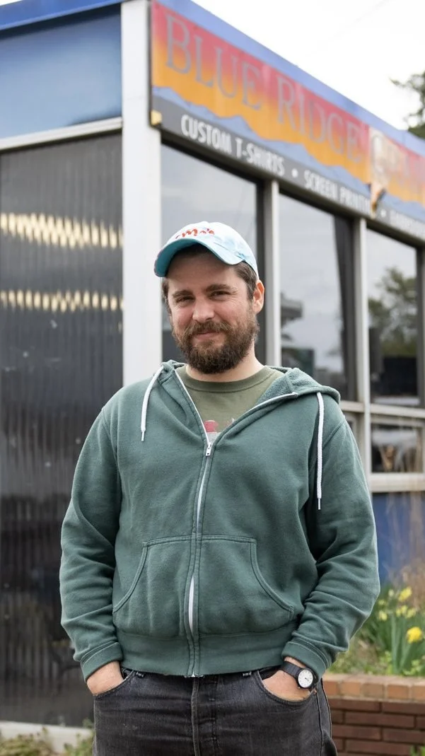 Man with beard wearing a green hoodie and a white baseball cap standing outside a storefront with a sign that reads "Blue Ridge".