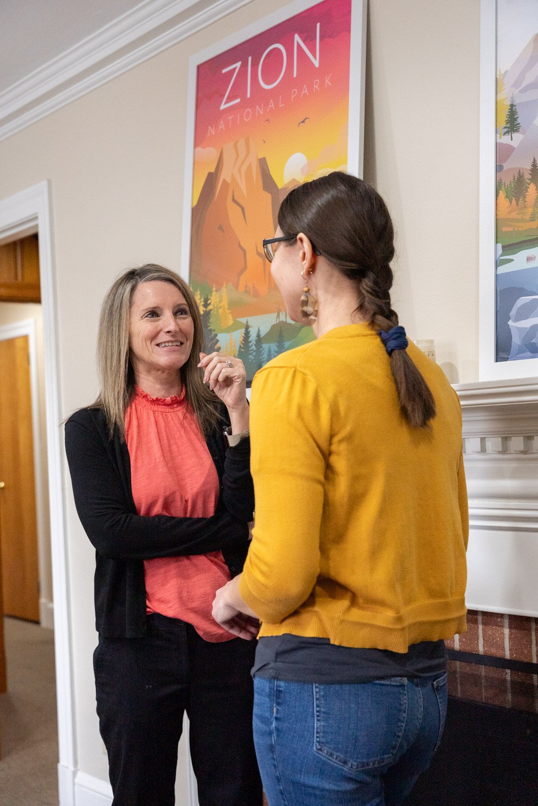 Two women having a conversation in front of a colorful Zion National Park poster inside a home or office.