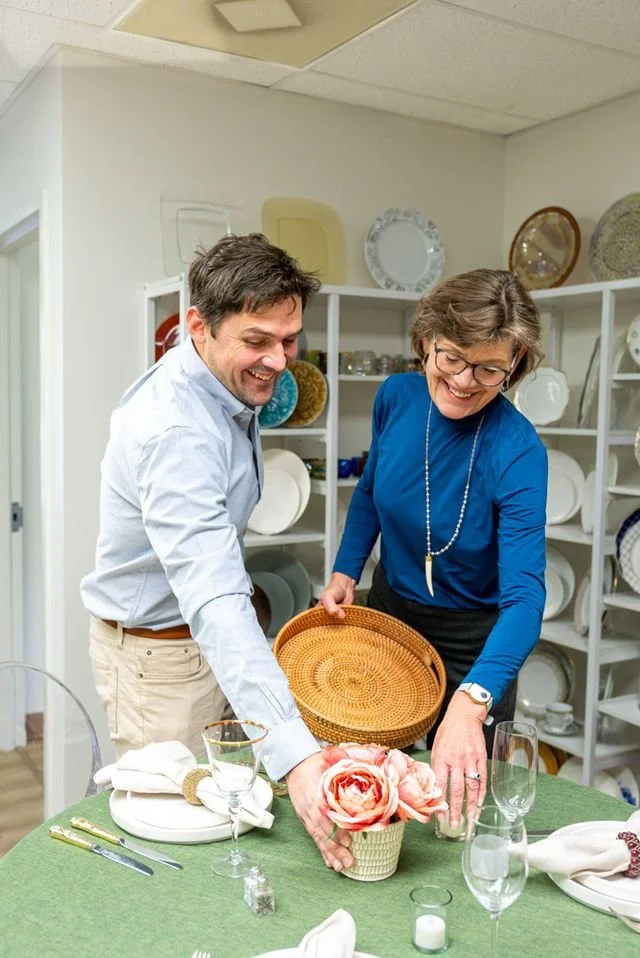 A man and a woman are setting a table with a green tablecloth, flowers, and glassware in a dining room with shelves of plates and bowls behind them.