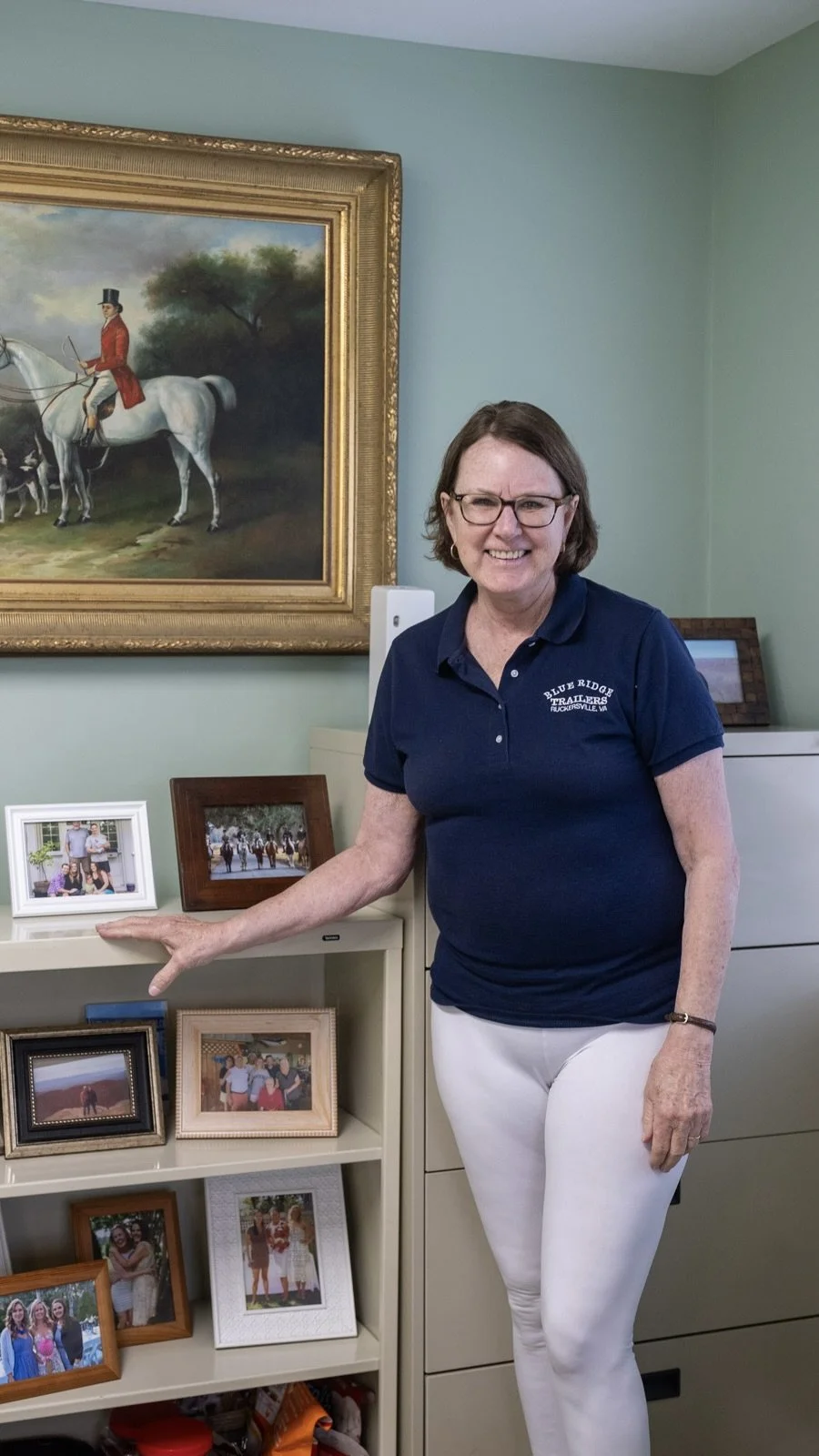 A smiling woman with glasses wearing a blue Polo shirt and white pants standing in a room with light green walls, pointing at a small framed photograph on a white shelf in front of her. Behind her, a large framed portrait of a man in a red coat riding a white horse hangs on the wall, and other framed photographs are on the shelves below.