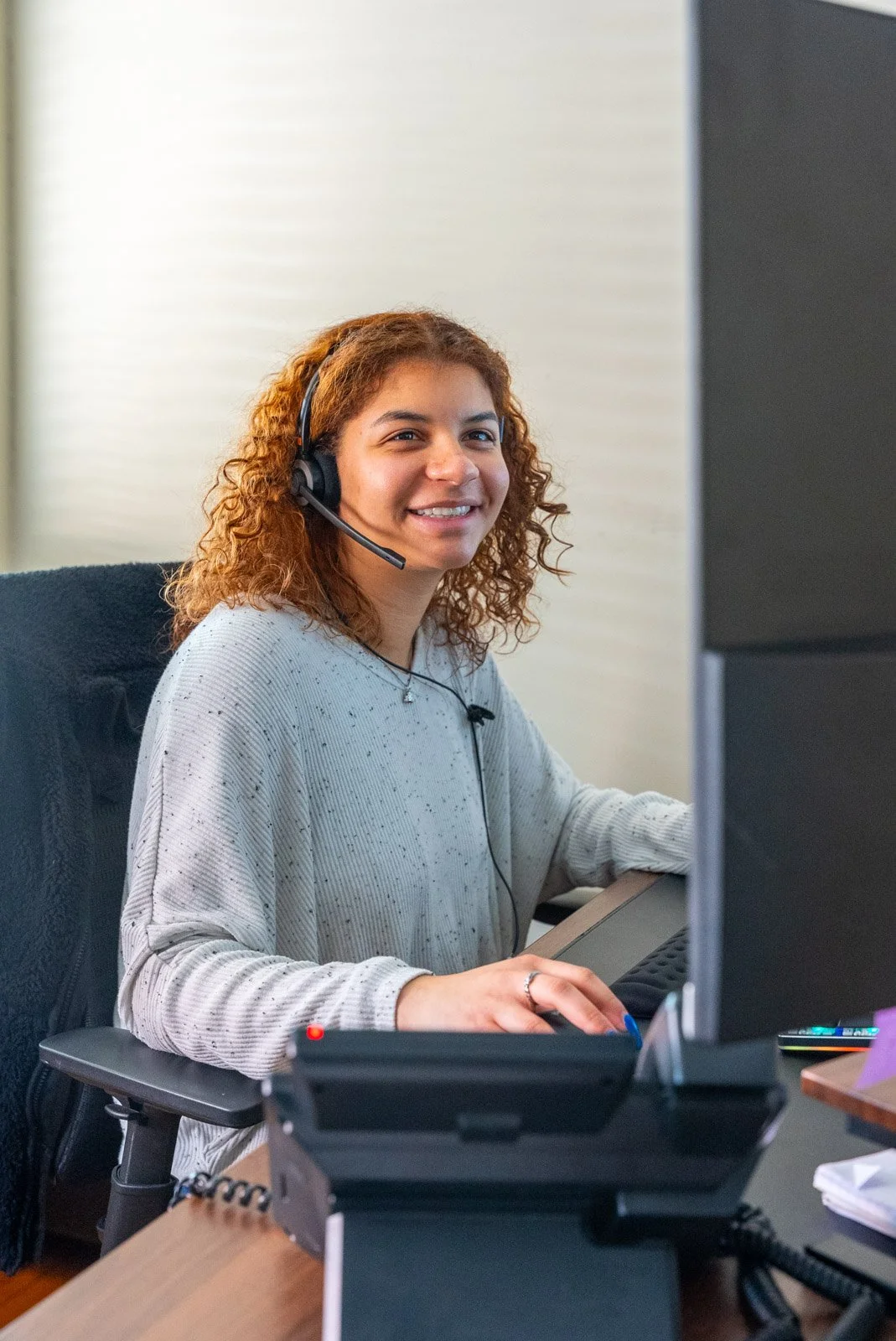 A woman with reddish curly hair wears a gray sweater and a headset, smiling while working at her desk in an office.