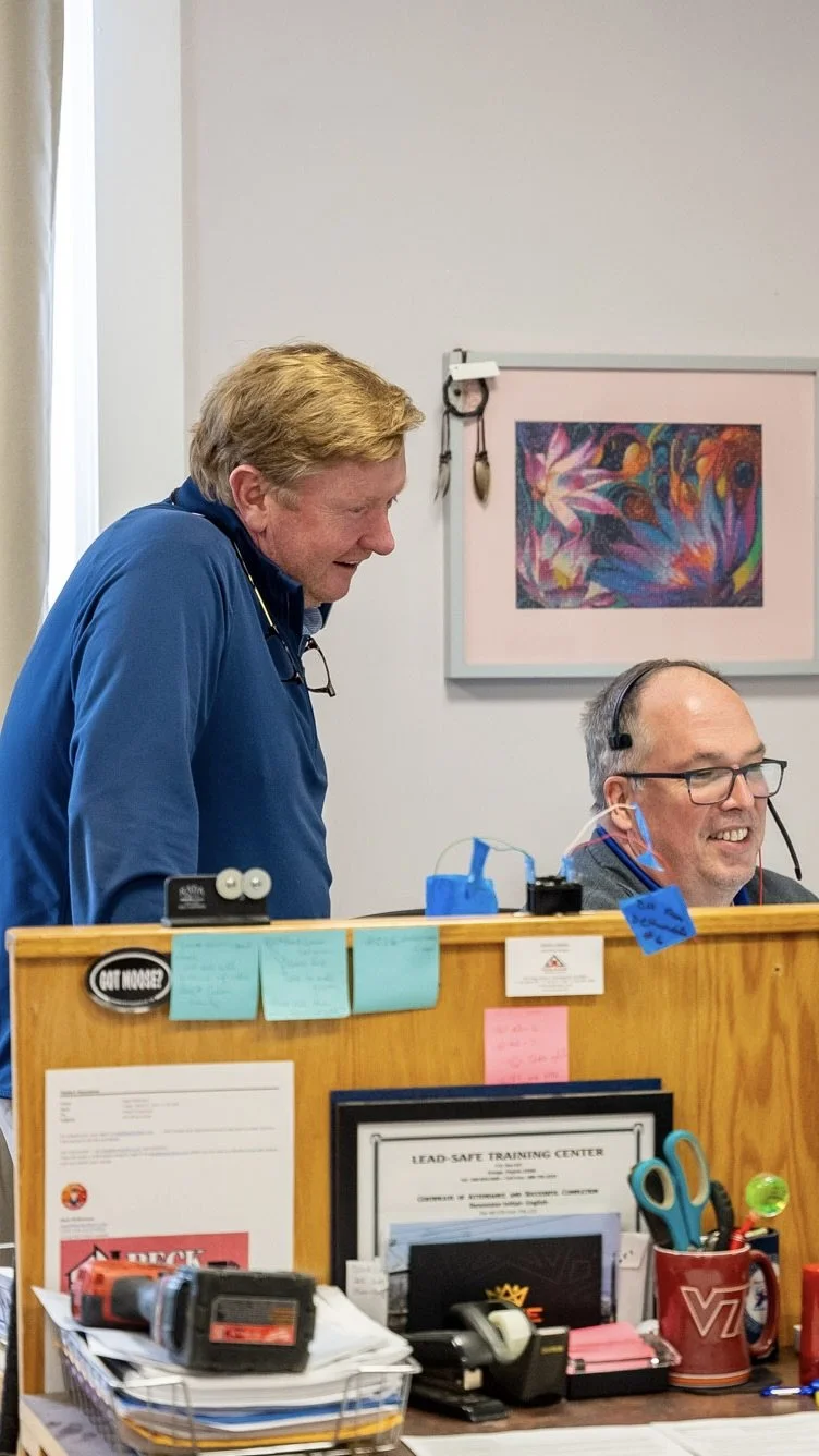 Two men working at an office reception desk, one standing and one sitting, with various office supplies and framed documents on the desk.