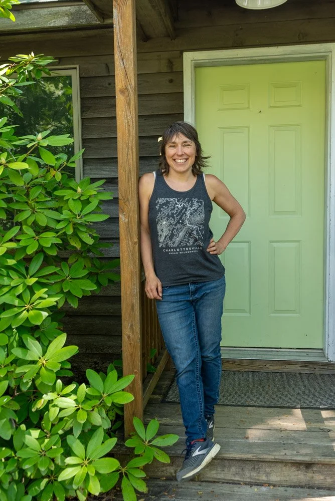 Woman standing on a wooden porch, smiling, wearing a black sleeveless shirt with graphic print, blue jeans, and sneakers, next to a green door and surrounded by green foliage.