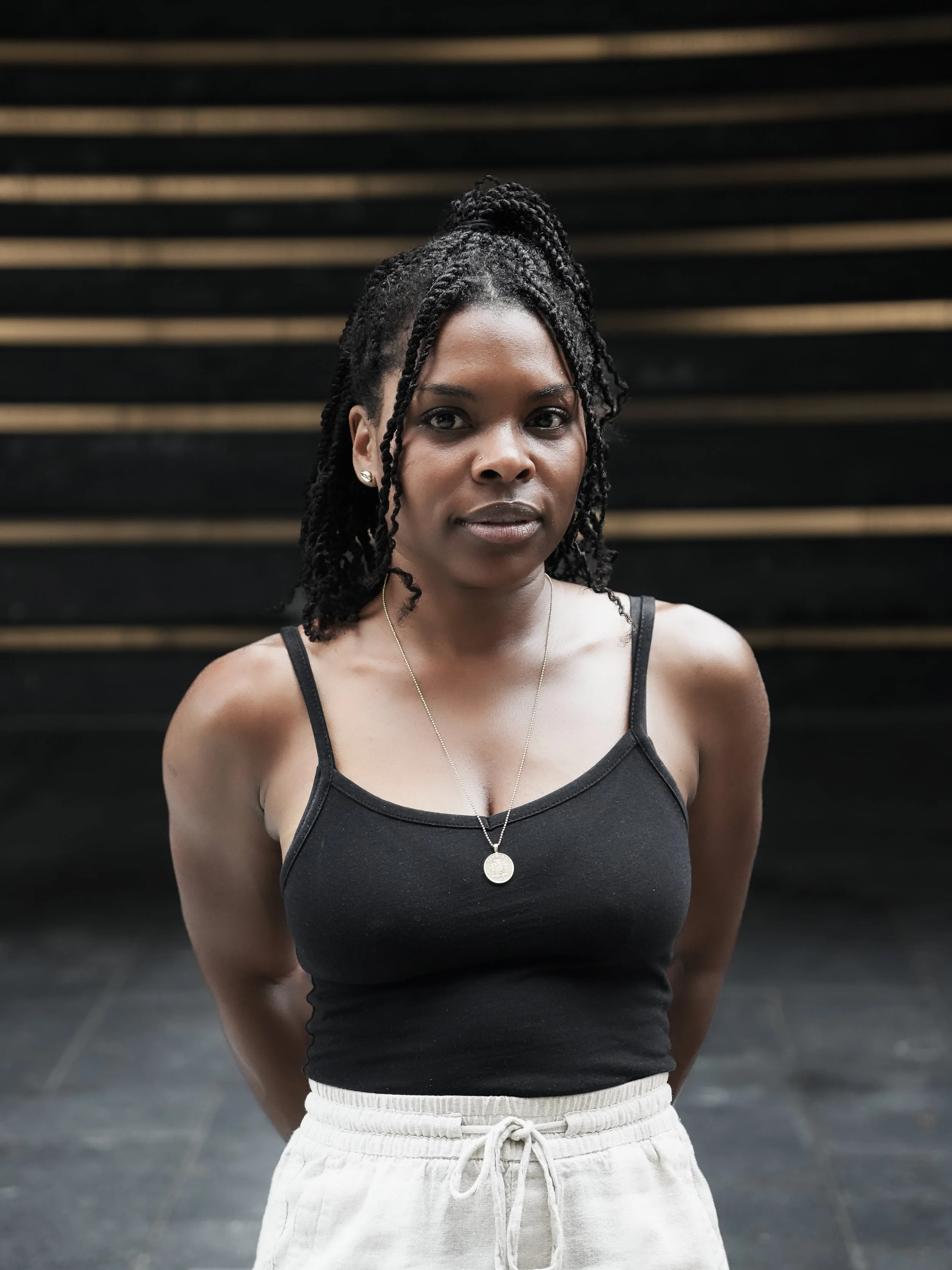 A woman with braided hair wearing a black tank top and beige drawstring pants, standing against a dark background with wooden steps.