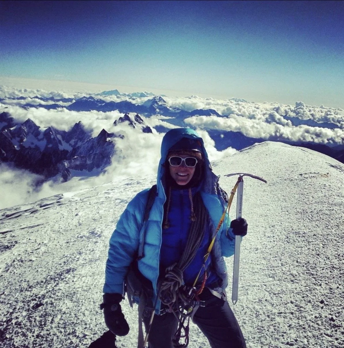A person in mountaineering gear standing on a snow-covered peak with a mountain range and clouds in the background.