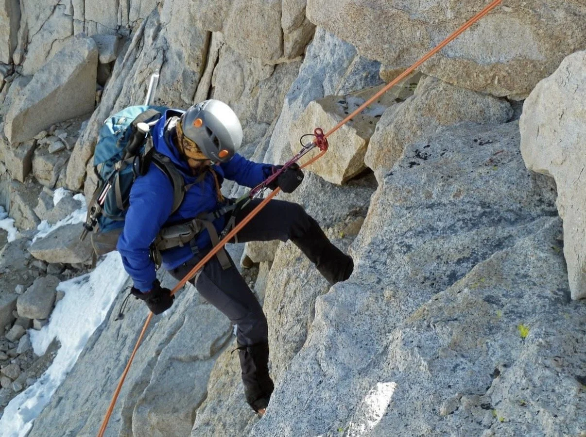 A person rock climbing on a steep rocky mountain face, wearing a helmet, harness, and backpack, holding onto an orange safety rope.