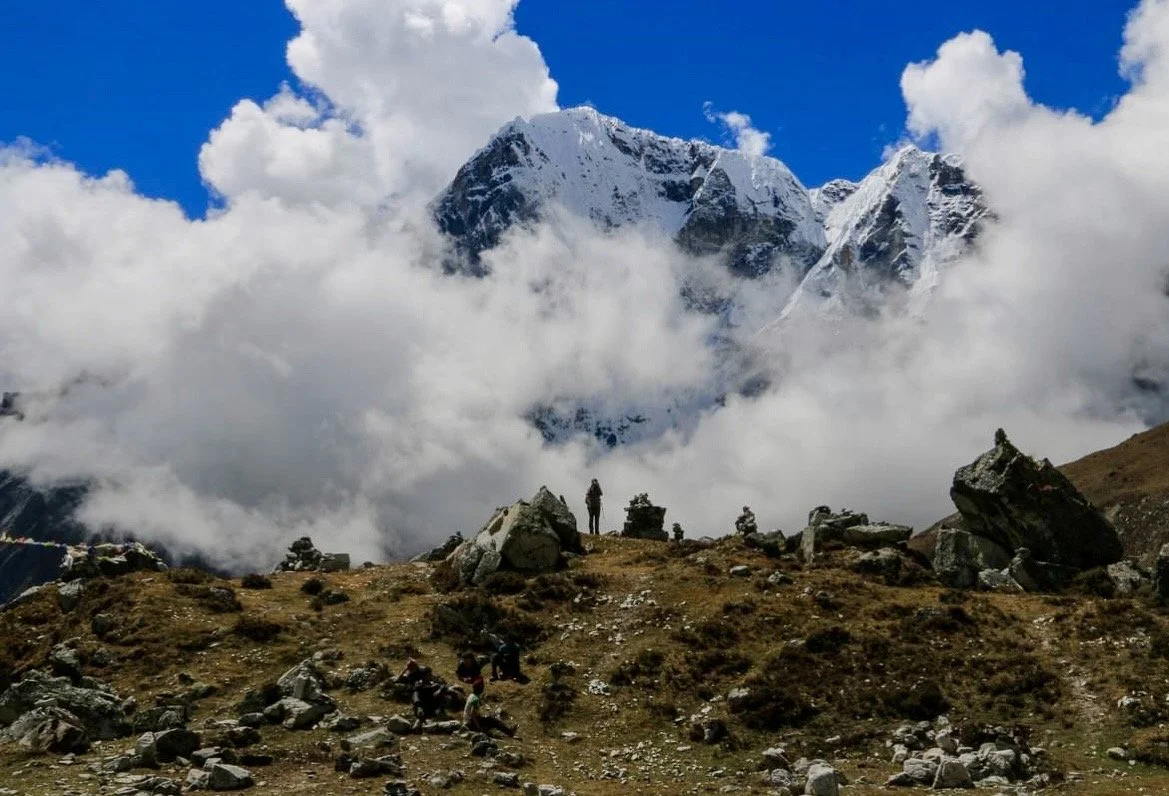 Mountain landscape with snow-capped peaks partially covered by clouds, rocky terrain in foreground, and a person standing on rocks.