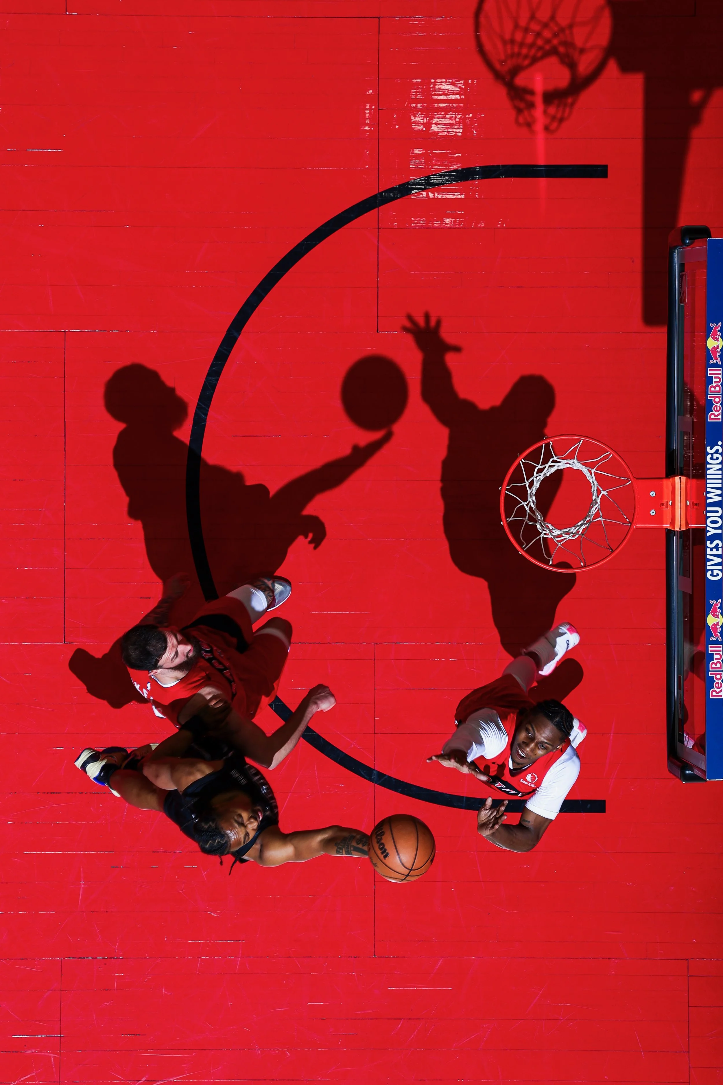 A basketball game viewed from above, showing players in red and black uniforms near the basket, with shadows cast on the red court, and a visible backboard with a net.