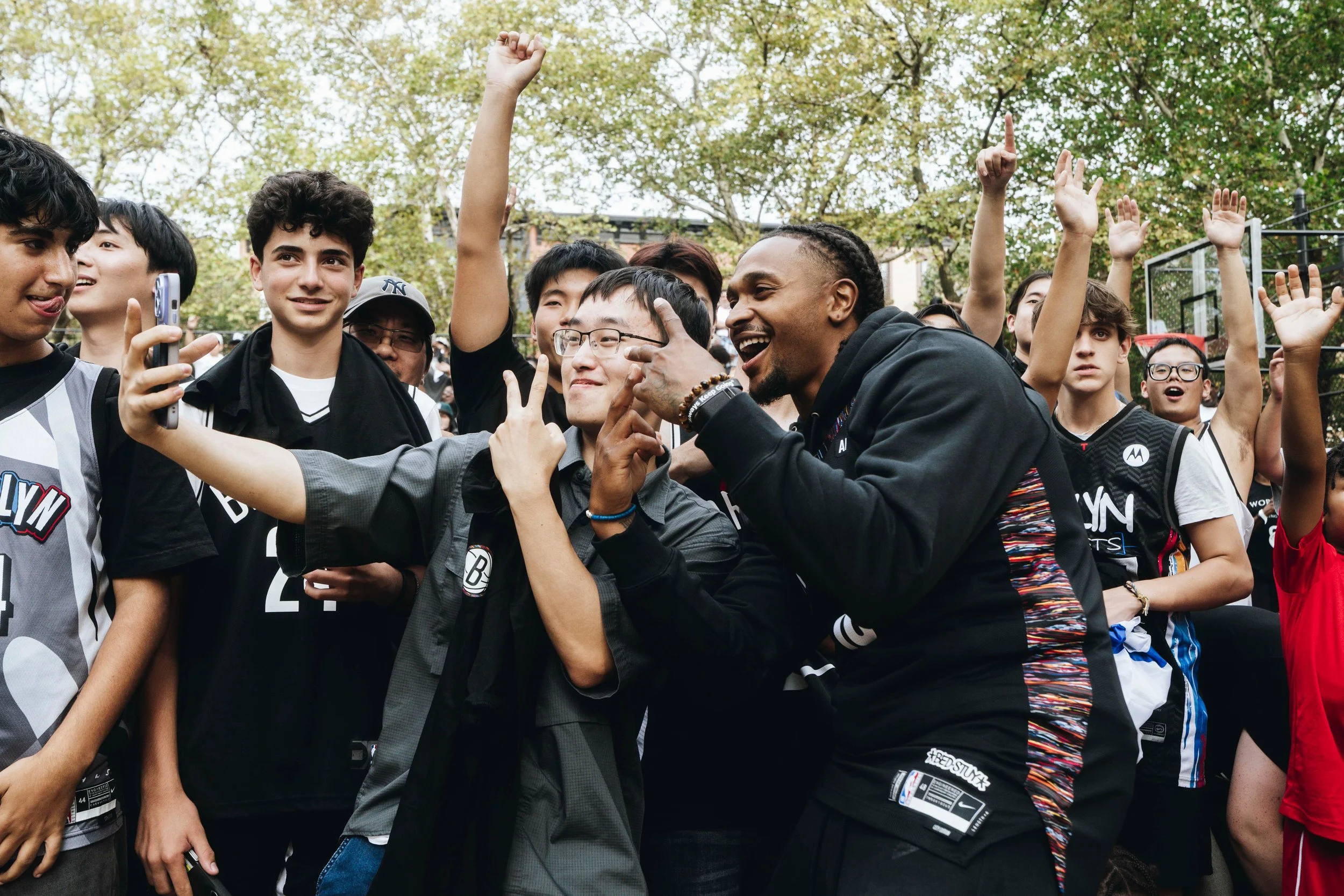 A diverse group of young people taking a selfie together outdoors, some raising their hands and smiling.