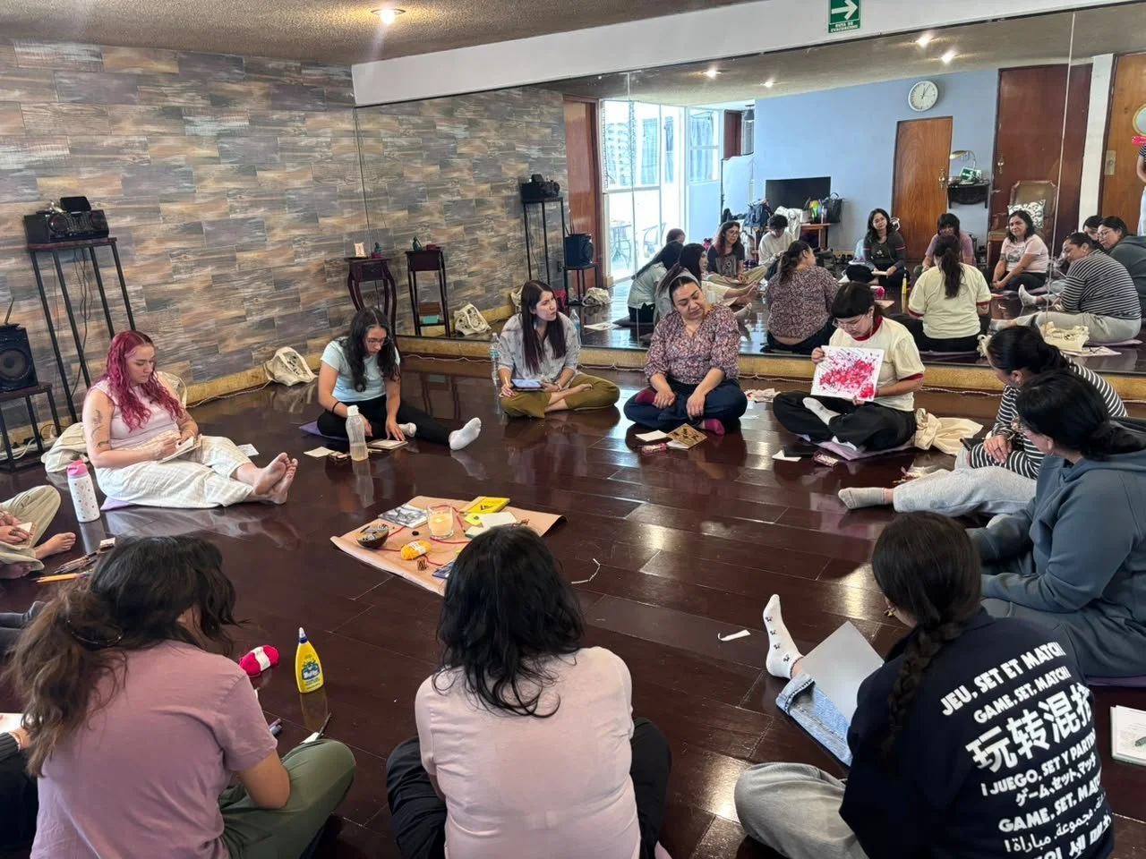 Grupo de personas sentadas en círculo en piso de madera en una habitación con pared de piedra y espejo grande, participando en una actividad grupal.
