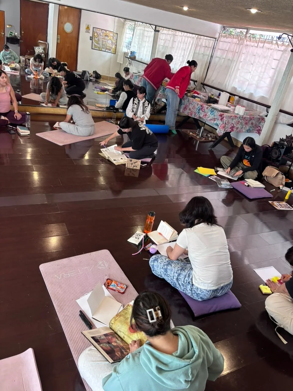 Grupo de personas sentadas en el suelo con colchonetas y libros, participando en una actividad en un espacio interior con mesas y ventanas.