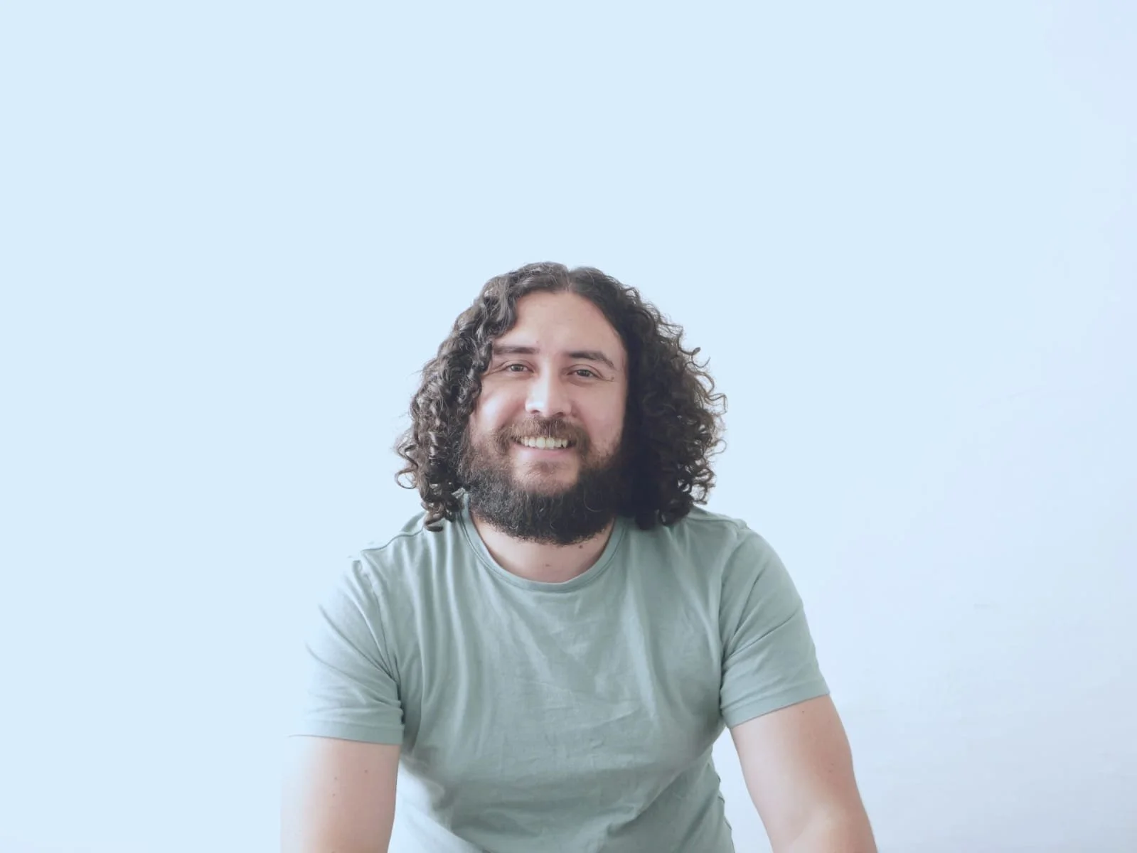 Hombre con cabello rizado, barba, camiseta de mezclilla y sonrisa, posando frente a una pared blanca.