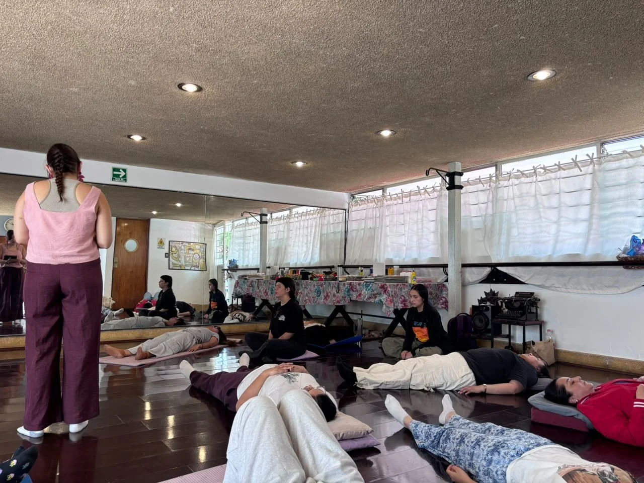 Grupo de personas descansando en una clase de yoga o meditación en un estudio con paredes de espejo y cortinas blancas.