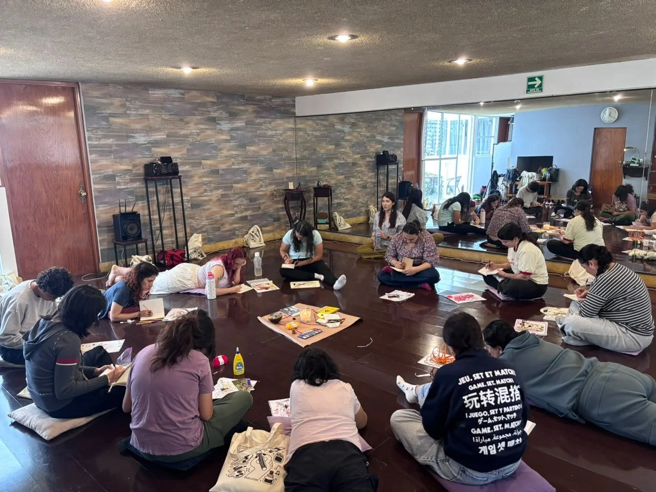Grupo de personas sentadas en el suelo, participando en una actividad de arte o manualidades en un salón interior.