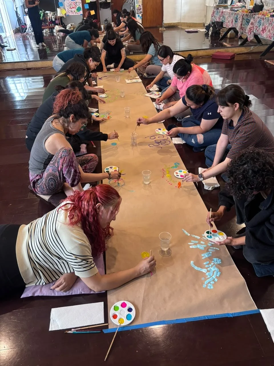 Personas participando en una actividad artística de pintura en una mesa larga en un salón cerrado.