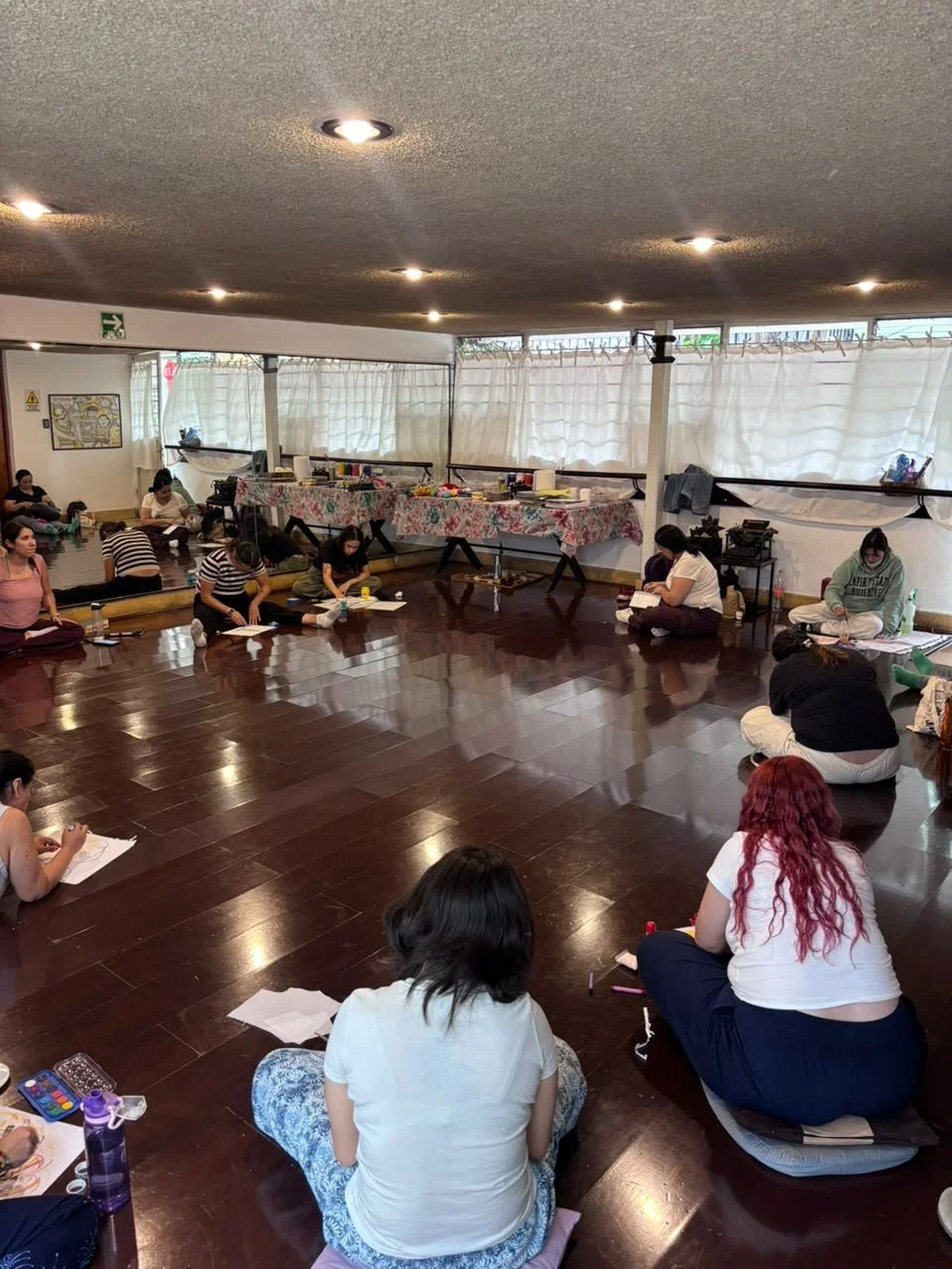 Personas sentadas en el suelo en un salón, participando en un taller o clase, con varias mesas con materiales en un fondo con cortinas blancas.