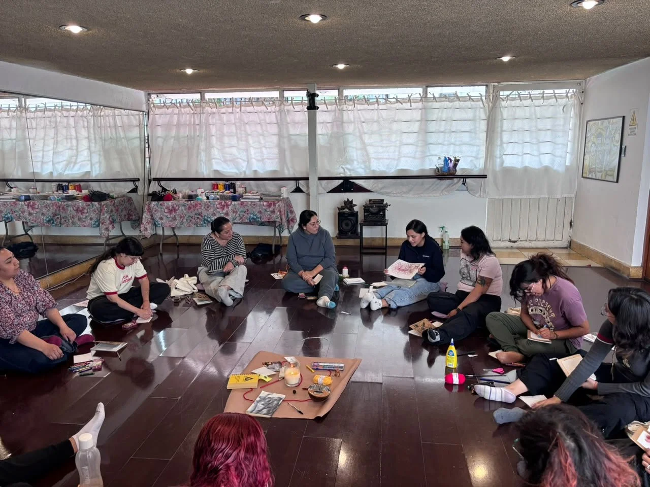 Un grupo de mujeres sentadas en círculo en una sala, participando en una actividad grupal, con mesas con objetos y materiales en el fondo.