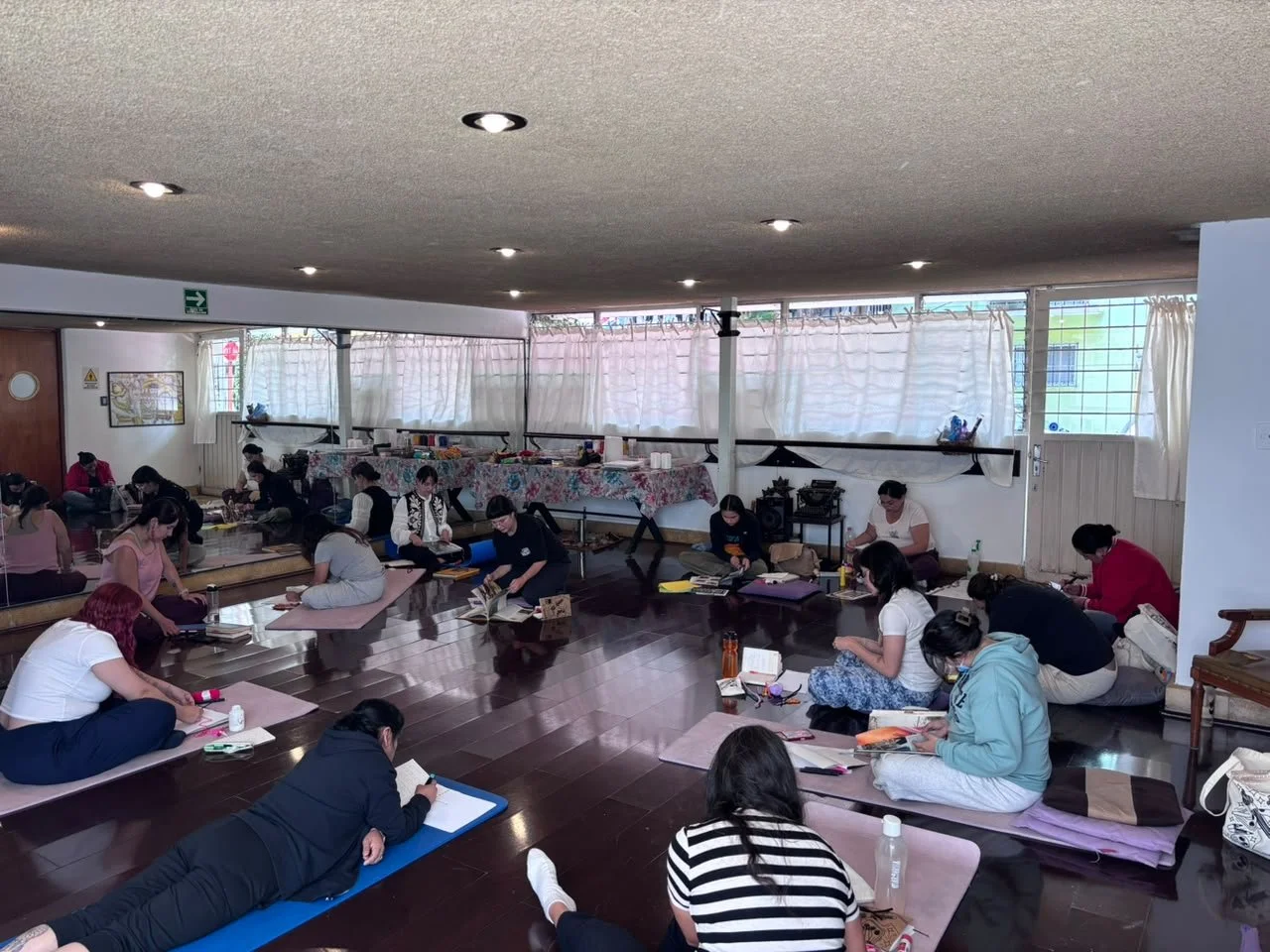 Grupo de personas en una clase o taller, sentadas en colchonetas, leyendo y tomando notas, en un espacio interior con ventanas con cortinas blancas.