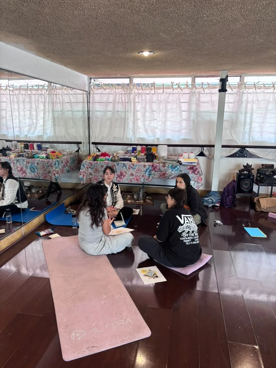 Cuatro personas sentadas en círculo en el suelo de una habitación con cortinas blancas, decoradas con manteles de flores, y objetos de maquillaje y accesorios en una mesa al fondo.
