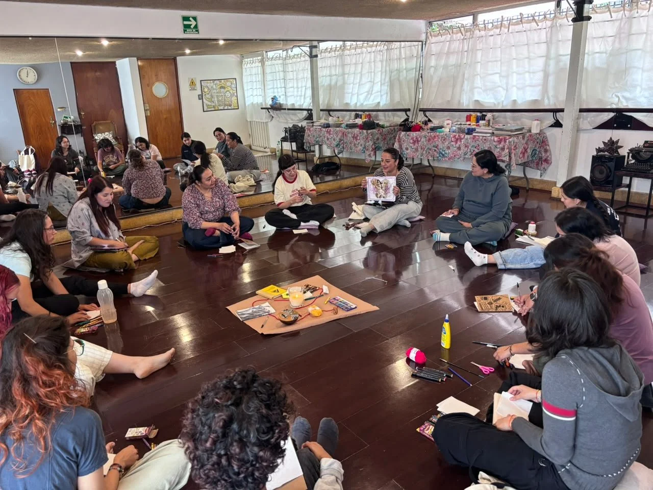 Grupo de personas sentadas en círculo en una sala, participando en un taller o reunión, con materiales dispersos en el centro.